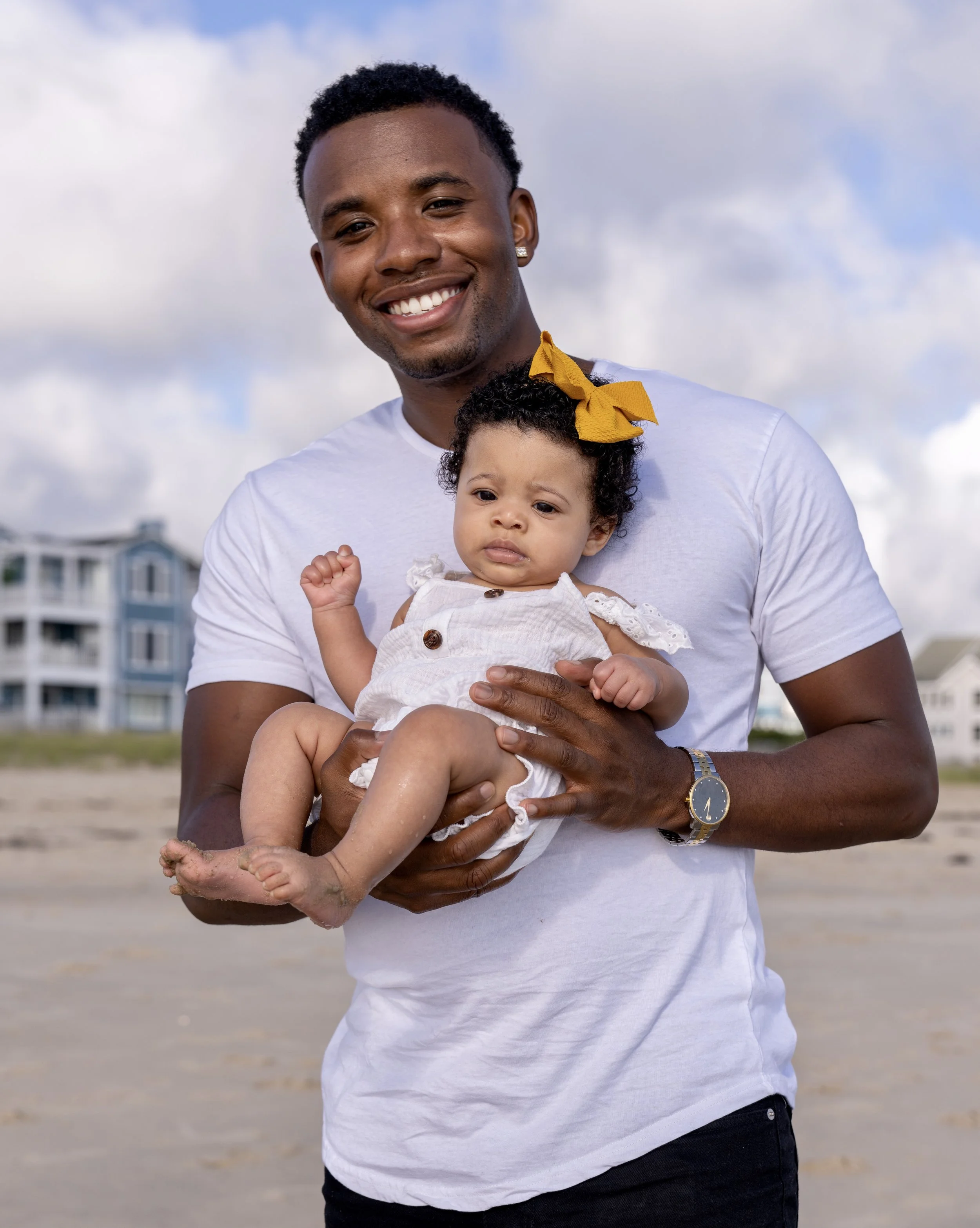 A man smiling and holding a young girl at the beach with buildings and a cloudy sky in the background.