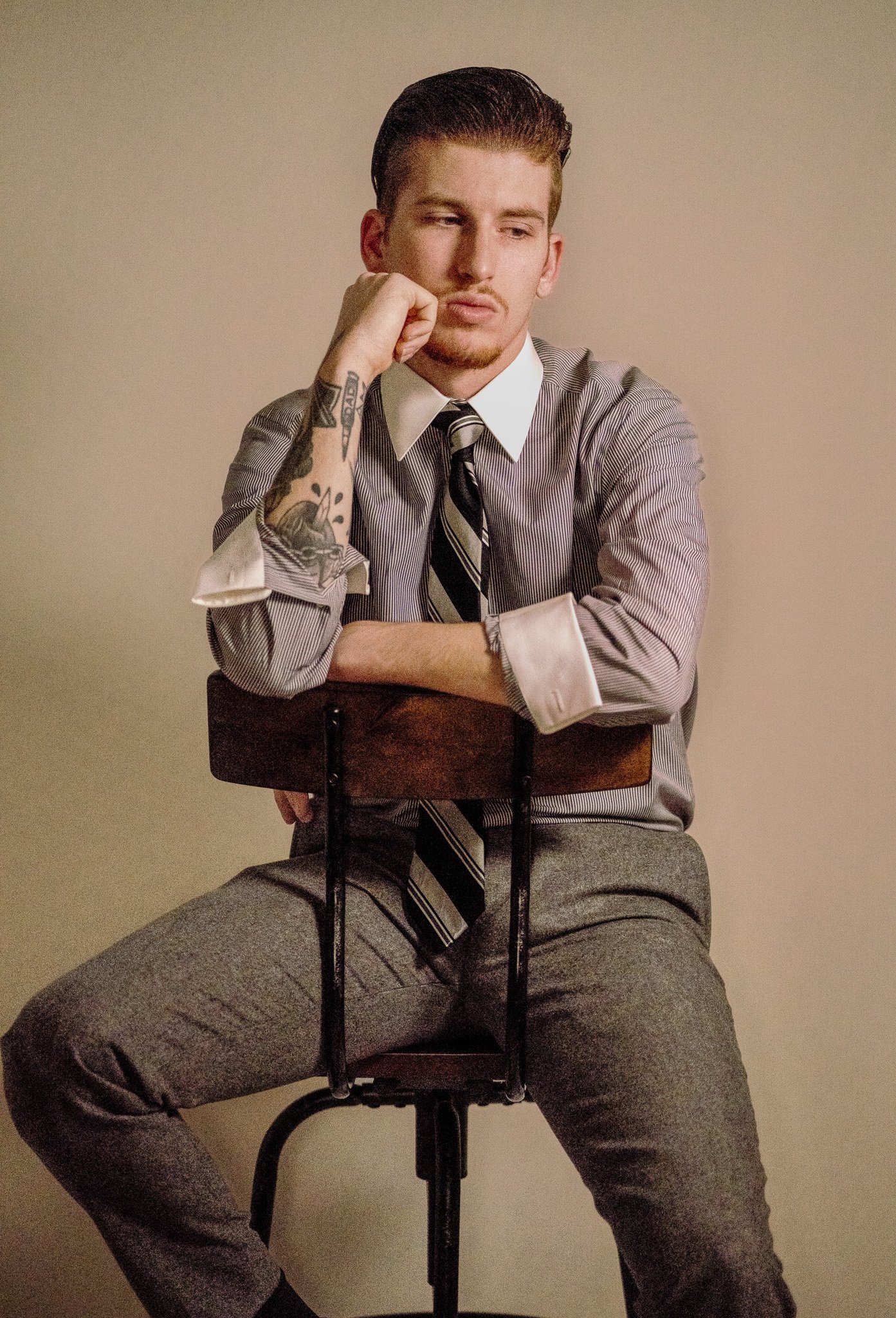 A young man with slicked back hair, facial hair, and tattoos on his left arm, wearing a striped dress shirt, tie, and gray pants, sitting on a chair with one leg over the other, resting his chin on his hand, against a plain beige background.
