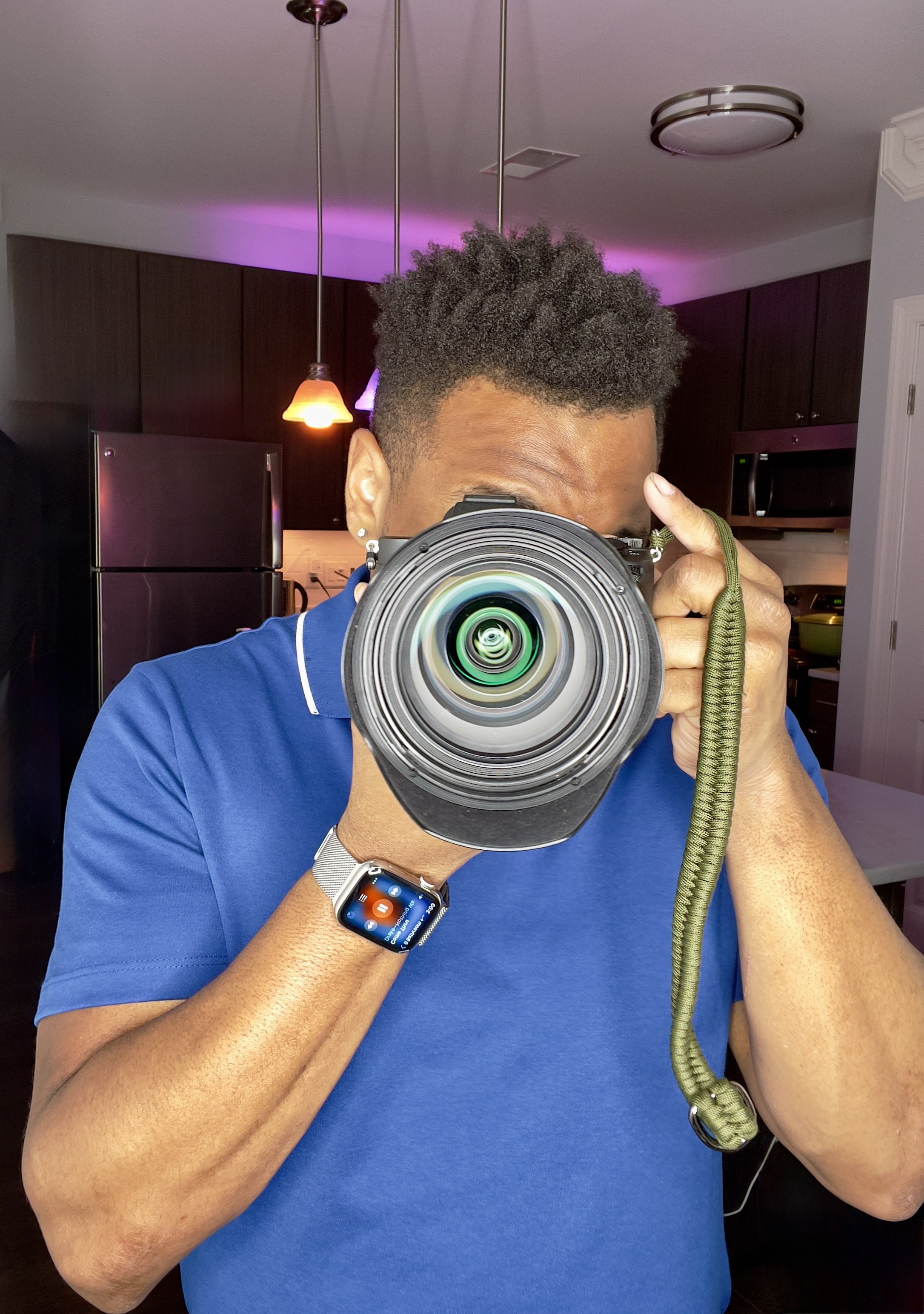 Person taking a selfie in a mirror with a camera, wearing a blue shirt and an Apple Watch, in a kitchen with dark cabinets and lighting.