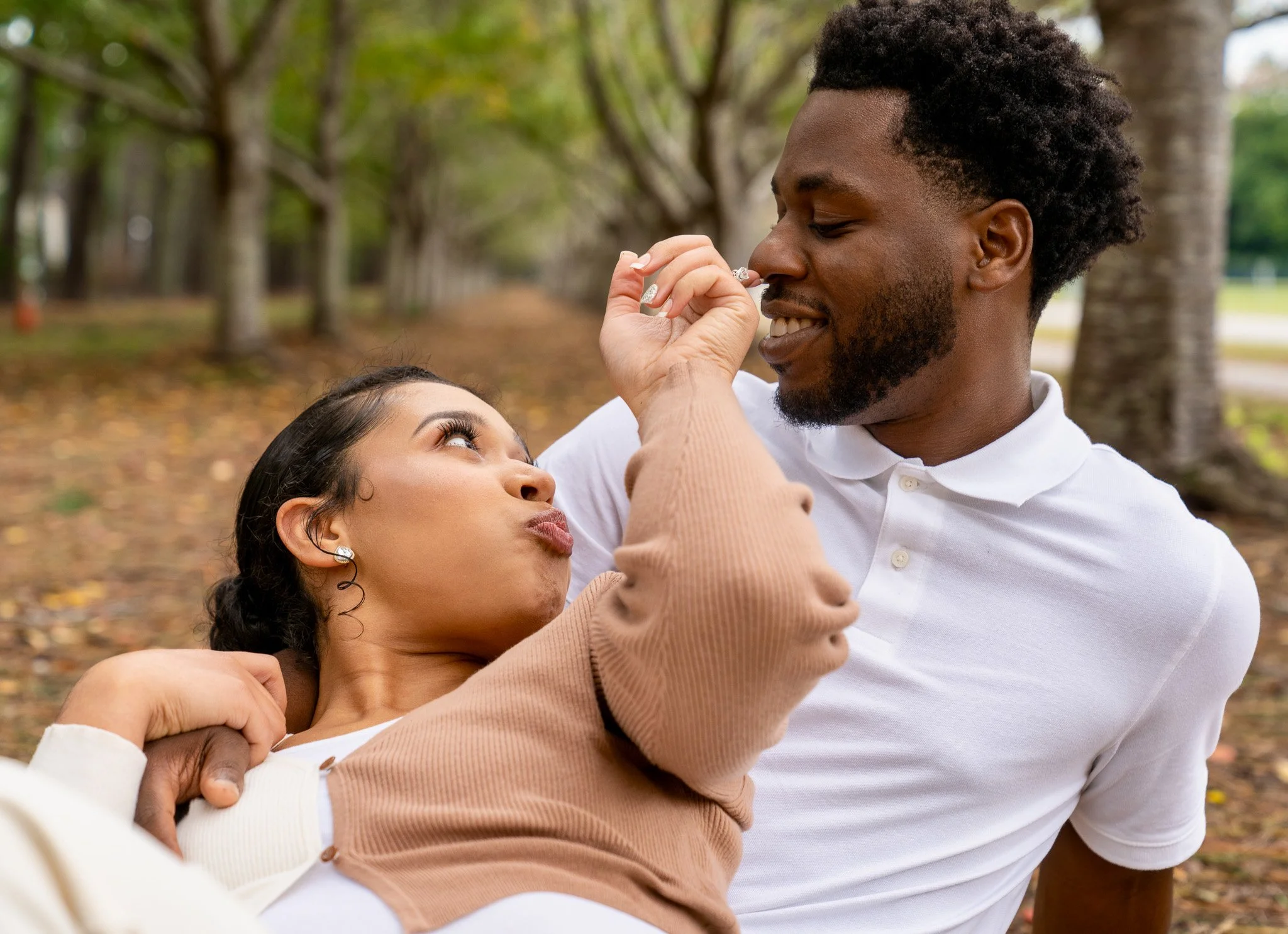 A man and woman are playfully holding each other's noses in a park with trees in the background.