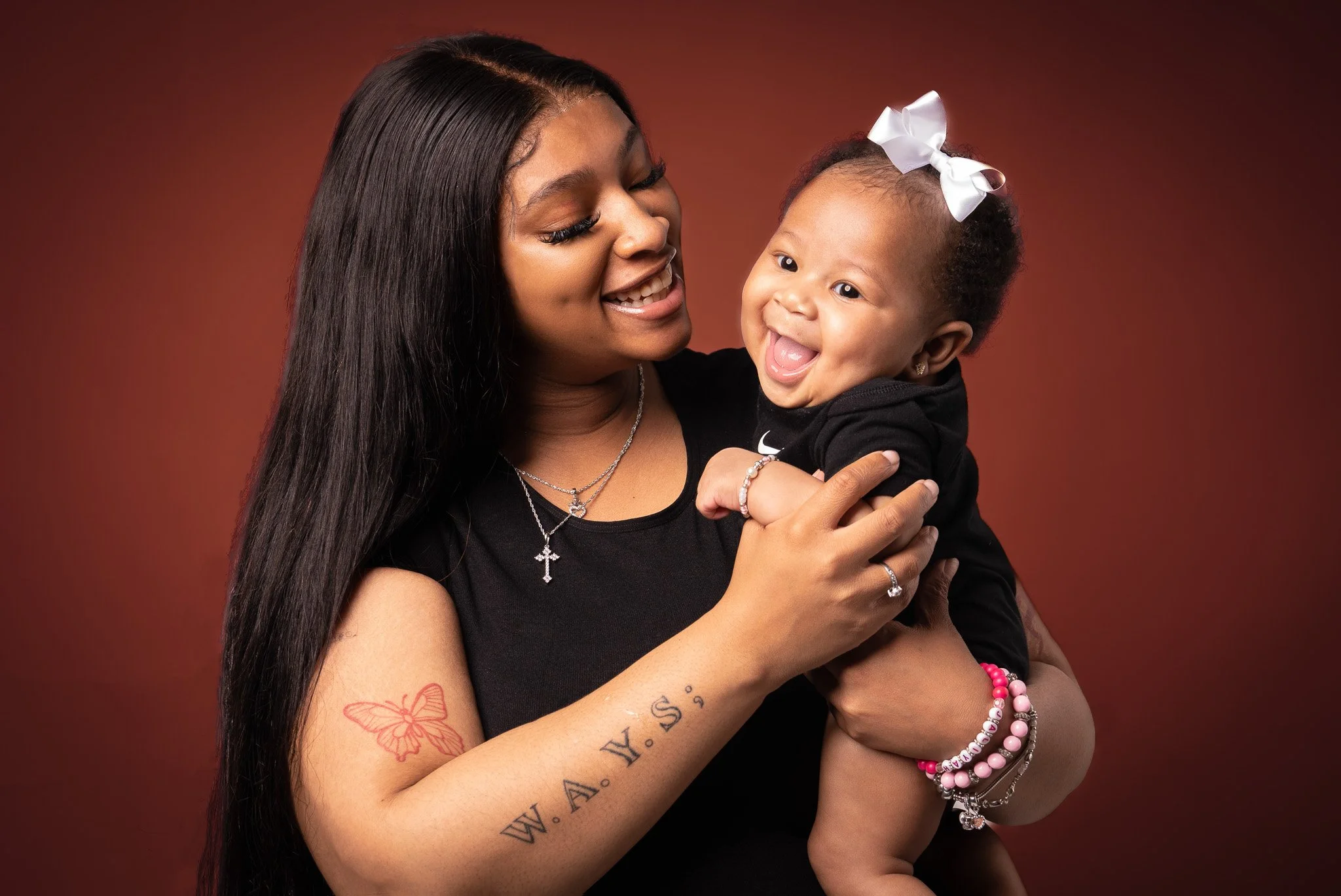 A woman with long black hair holding a smiling young girl with curly hair and a white bow in her hair, against a plain red background.