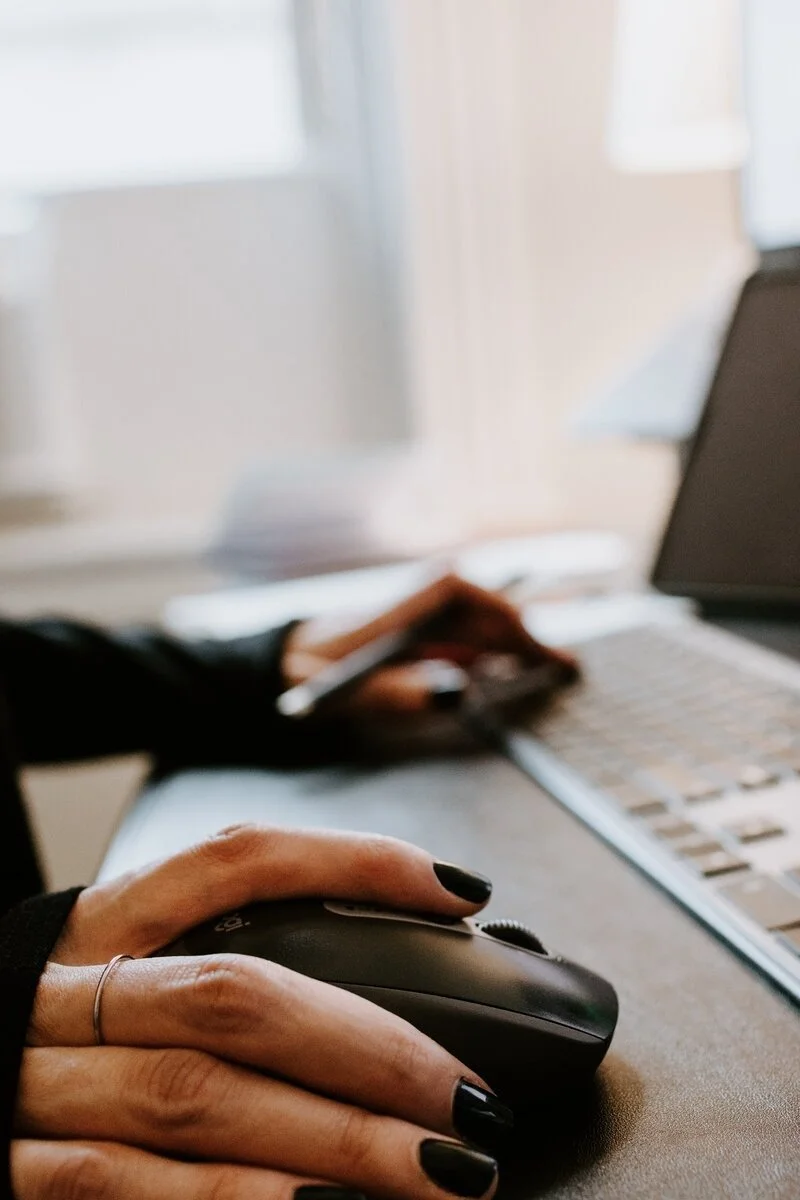 Close-up of a person's hand with black painted nails using a computer mouse on a desk with a laptop and smartphone in the background.