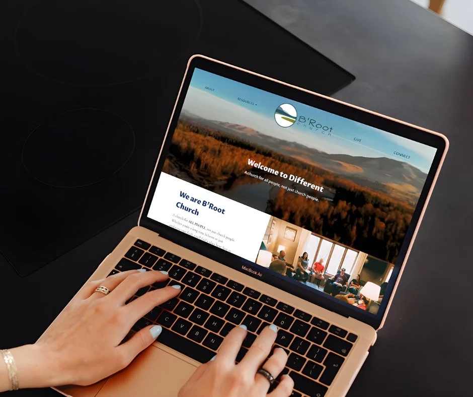 A person using a rose gold MacBook Air laptop on a black surface, browsing the B'Root Church website with a scenic landscape in the header.