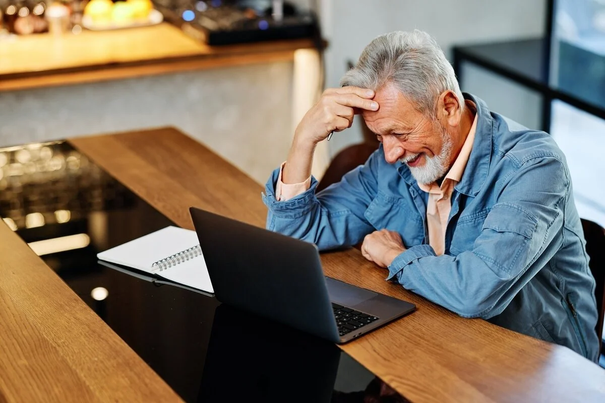 An older man with gray hair and a beard, smiling and holding his forehead, looking at a laptop screen in front of him.
