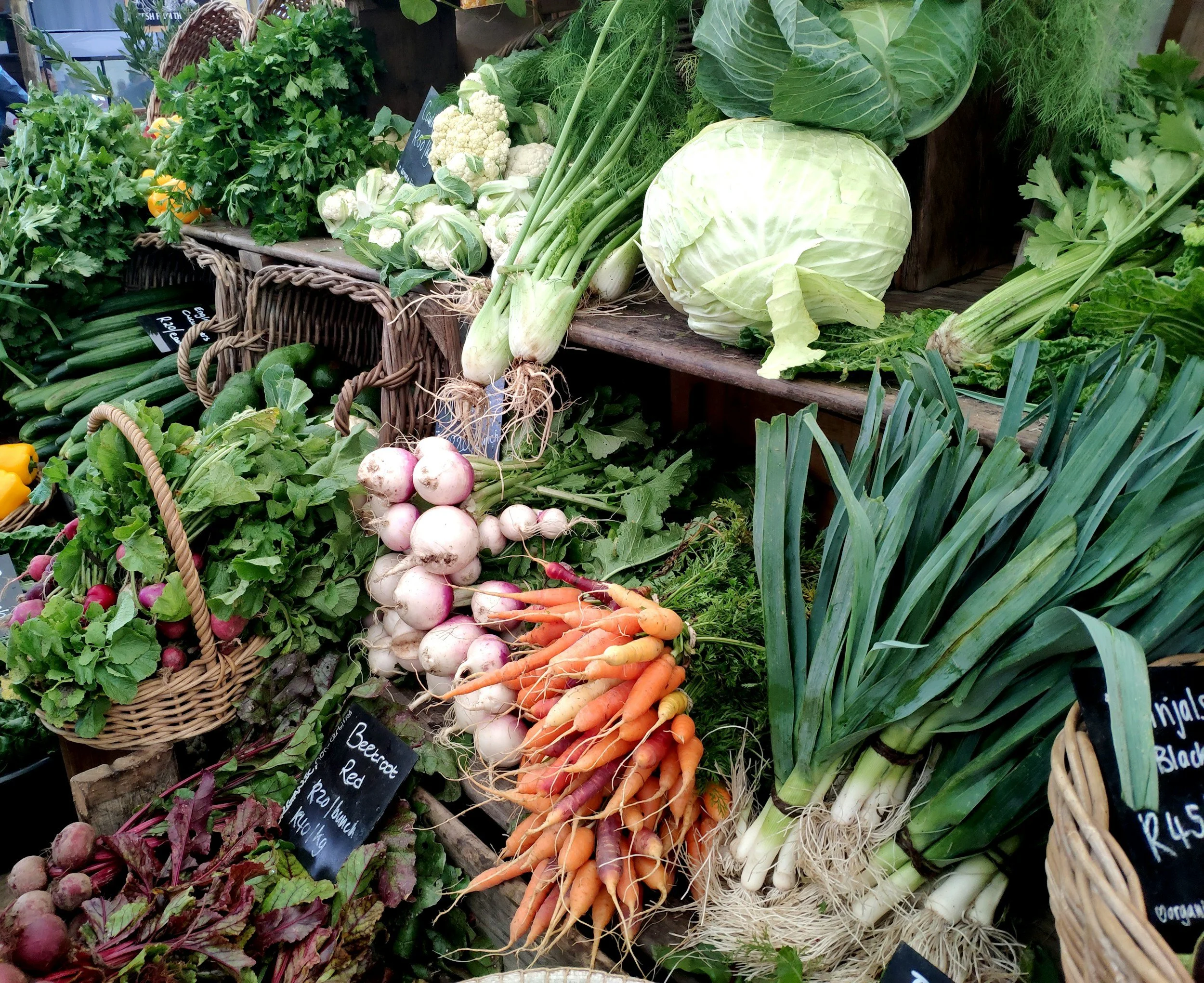 A display of fresh vegetables at a farmer's market, including cauliflower, cabbage, carrots, turnips, radishes, green onions, zucchinis, and leafy greens, arranged in baskets and on wooden shelves.