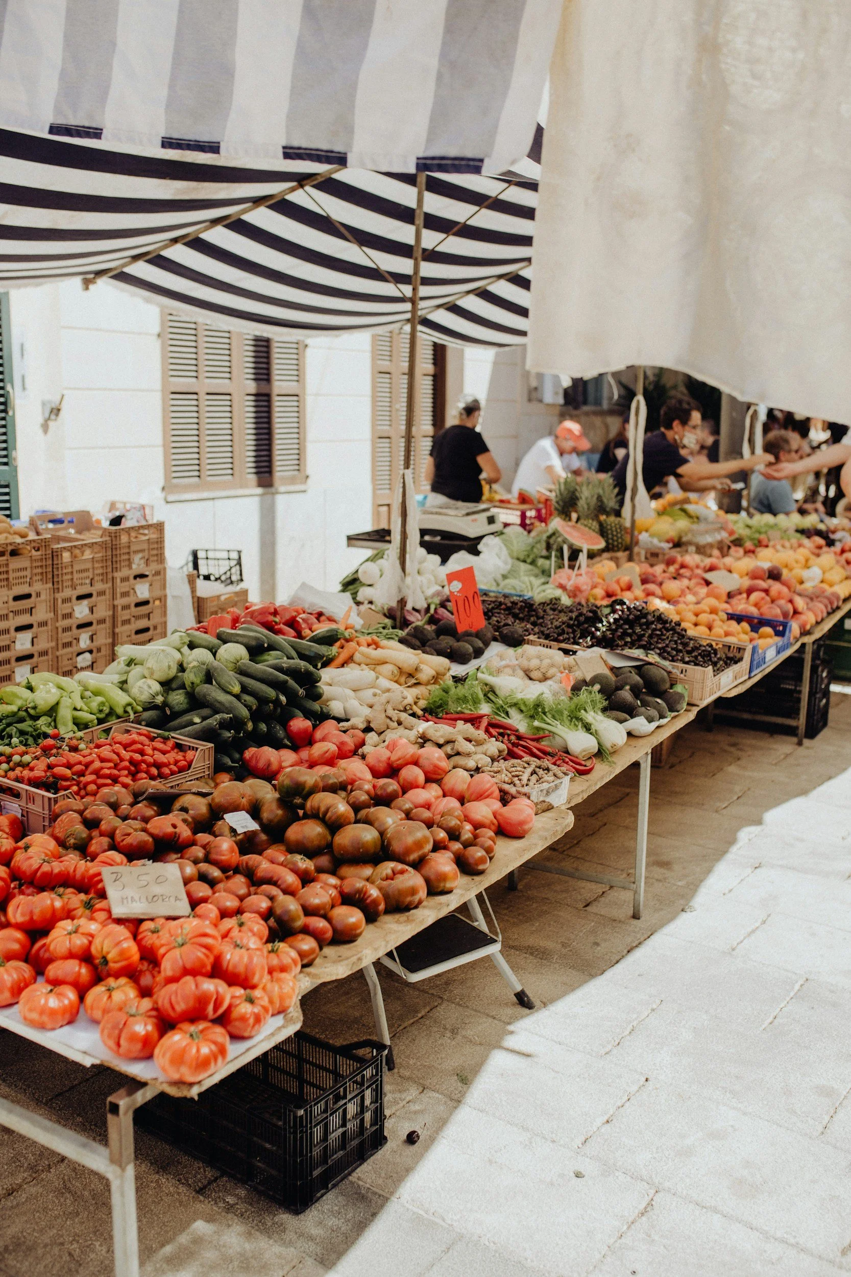 Outdoor market stall displaying a variety of fresh vegetables, including tomatoes, cucumbers, and peppers, with a striped canopy overhead and people shopping in the background.