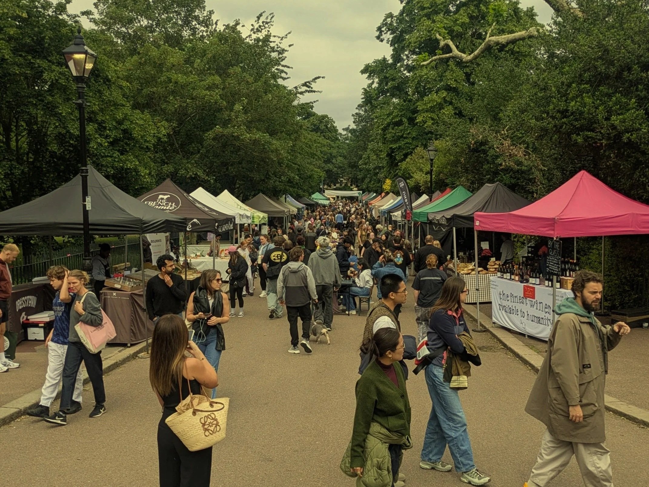 Crowd of people at an outdoor marketplace or fair with tents and trees in the background.