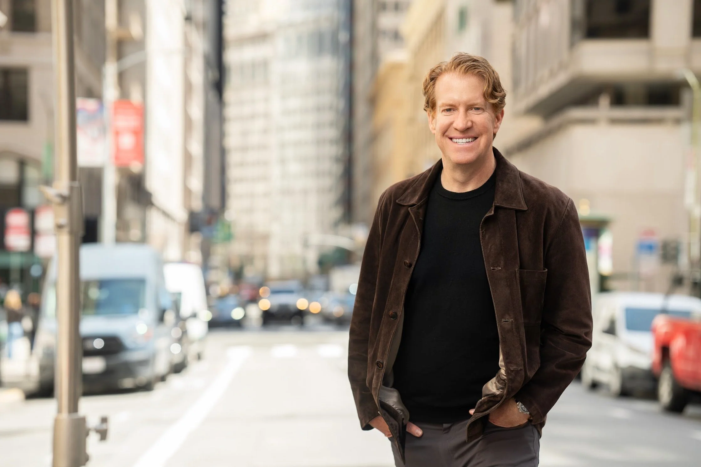 A smiling man with curly blonde hair stands on a city street, wearing a brown jacket over a black shirt, with cars and tall buildings in the background.