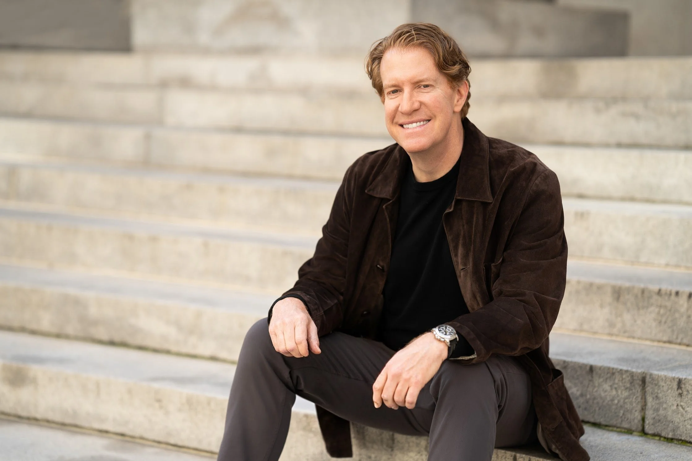 A middle-aged man with beige hair, smiling, sitting on concrete stairs outside, wearing a brown jacket, black shirt, and gray pants.