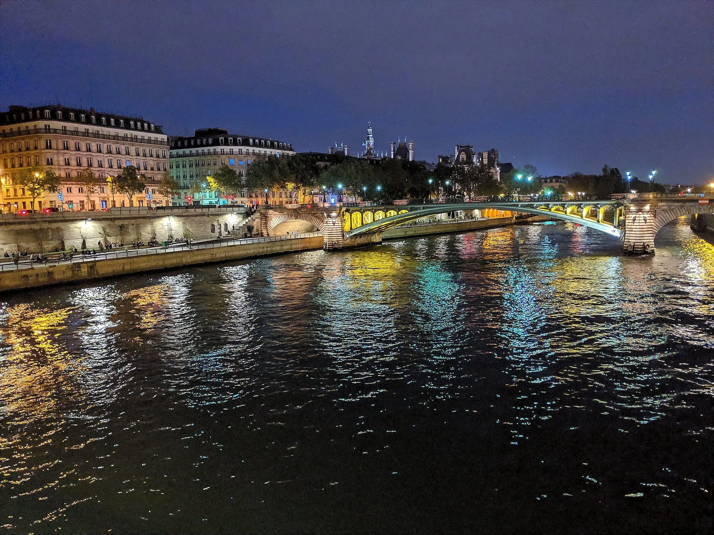 Nighttime view of a cityscape with illuminated buildings, a bridge over a river, and reflections of lights on the water.