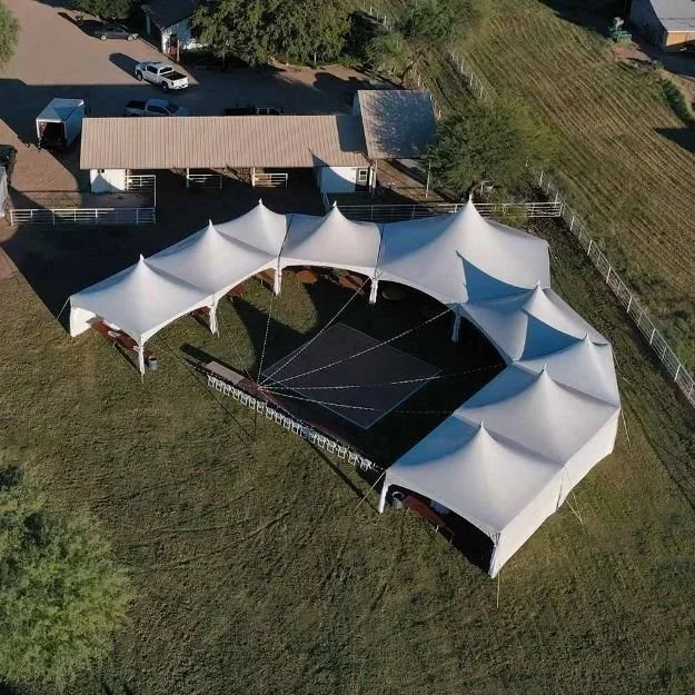 Aerial view of a large outdoor event tent set up on grassy area, surrounded by a fence and adjacent to a building with parked cars nearby.