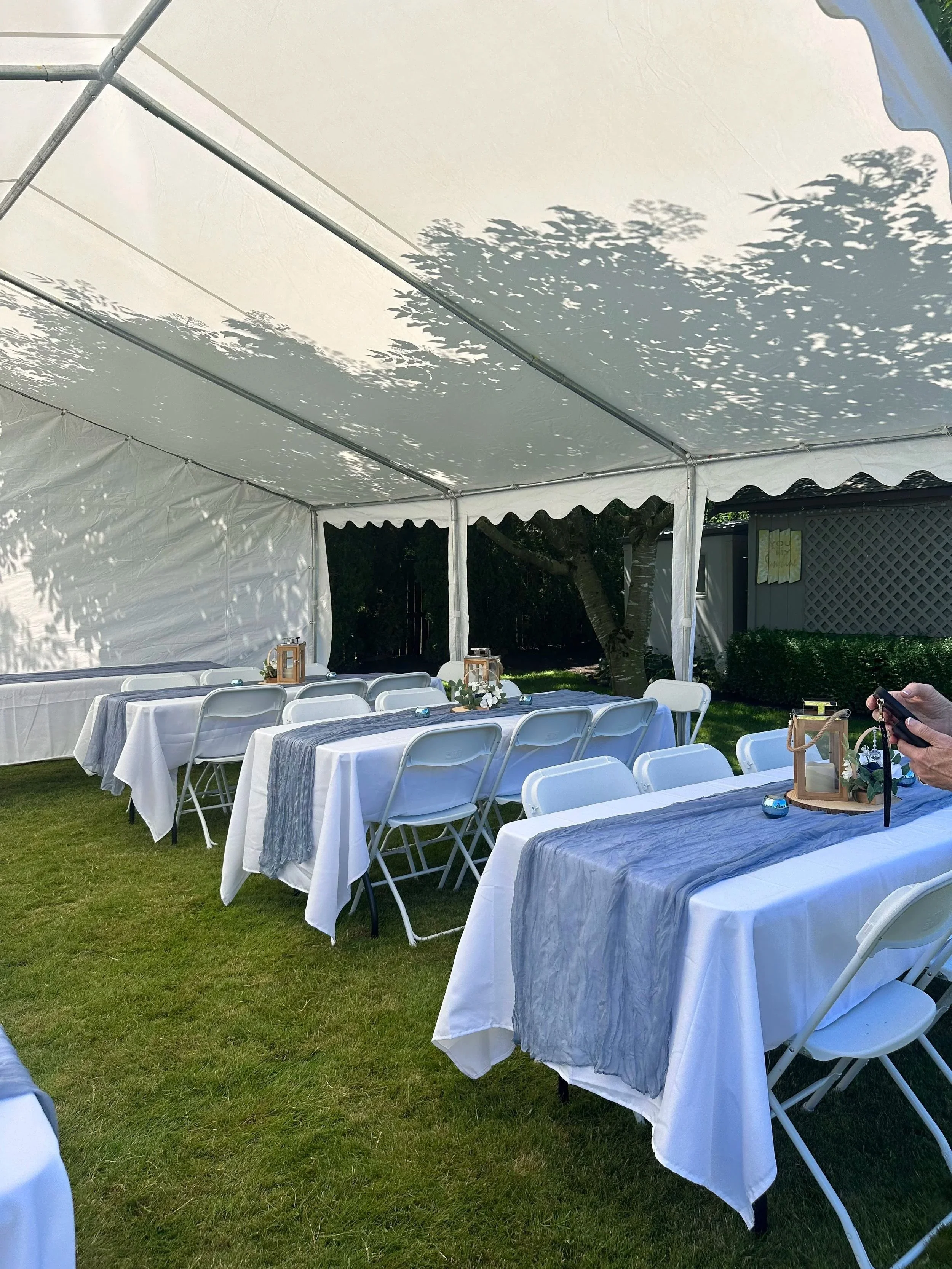 An outdoor event setup with long tables covered in white tablecloths and gray runners, surrounded by white folding chairs, under a white marquee tent, with decorative lanterns, flower arrangements, and small candles, on a grassy lawn with trees in th