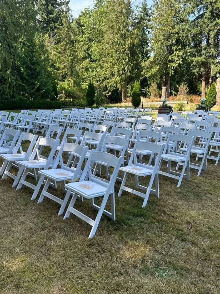 Rows of white folding chairs arranged outdoors on grass, facing a garden with large trees and shrubs, likely set up for an outdoor event or wedding.