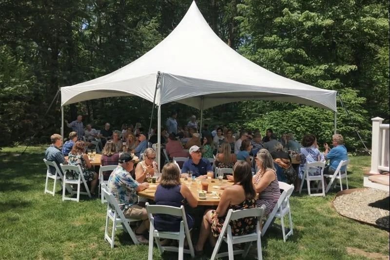 Gathering of people sitting around tables under a white canopy tent outdoors, surrounded by green trees.