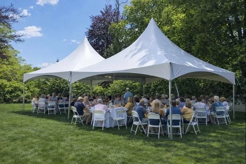 Outdoor gathering under a large white tent with multiple tables and chairs on a grassy lawn surrounded by trees.