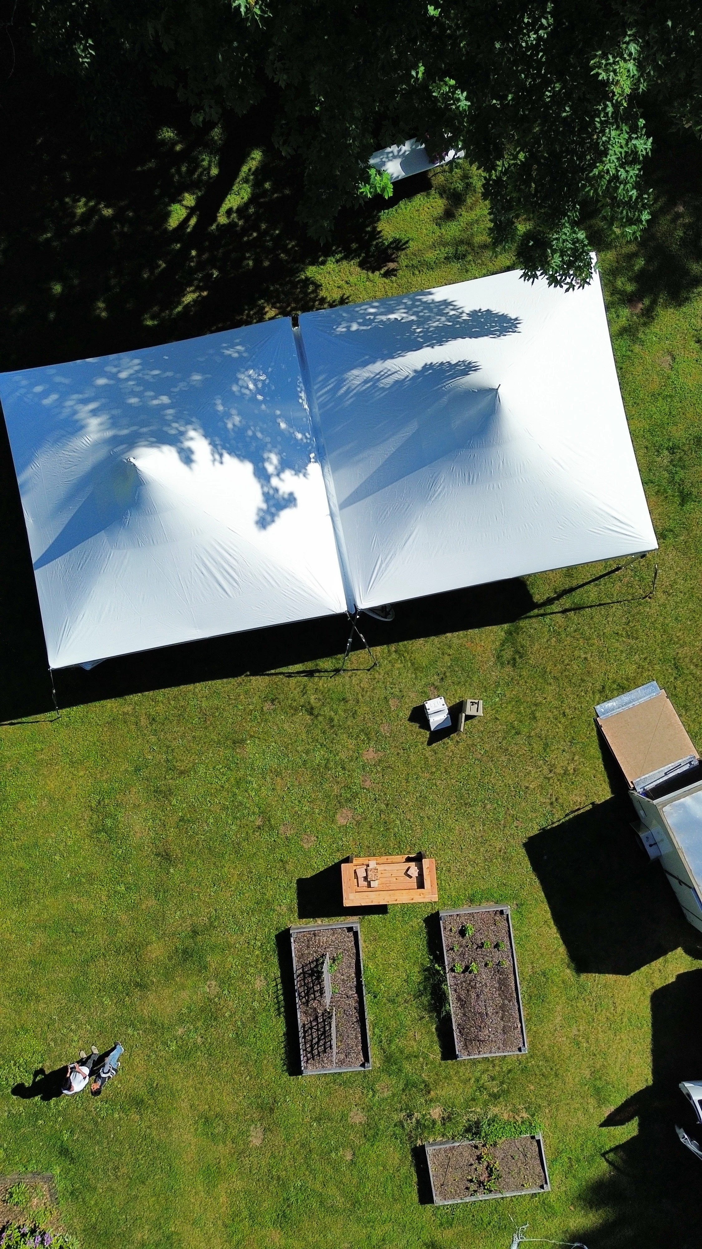 An aerial view of a backyard with a large white outdoor canopy, garden beds, and a few people sitting on the grass.