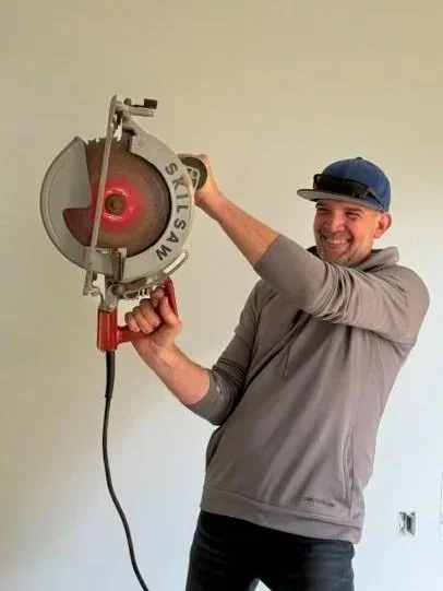 Man wearing blue baseball cap and sweatshirt, smiling, and holding up a large saw.