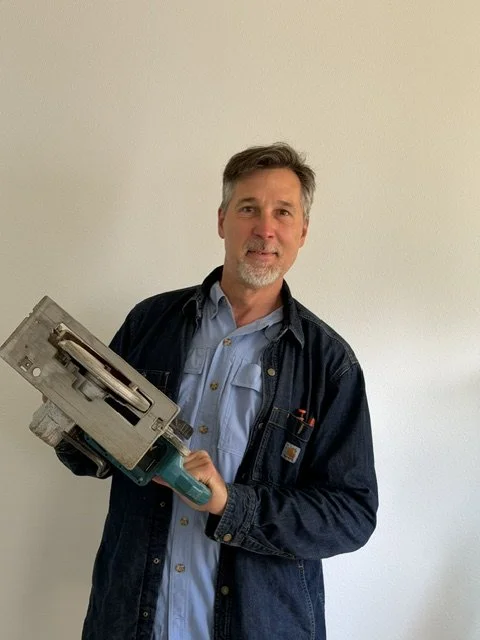 Tall man with grey beard and brown hair, wearing a denim button up and clue jacket, posing with a saw