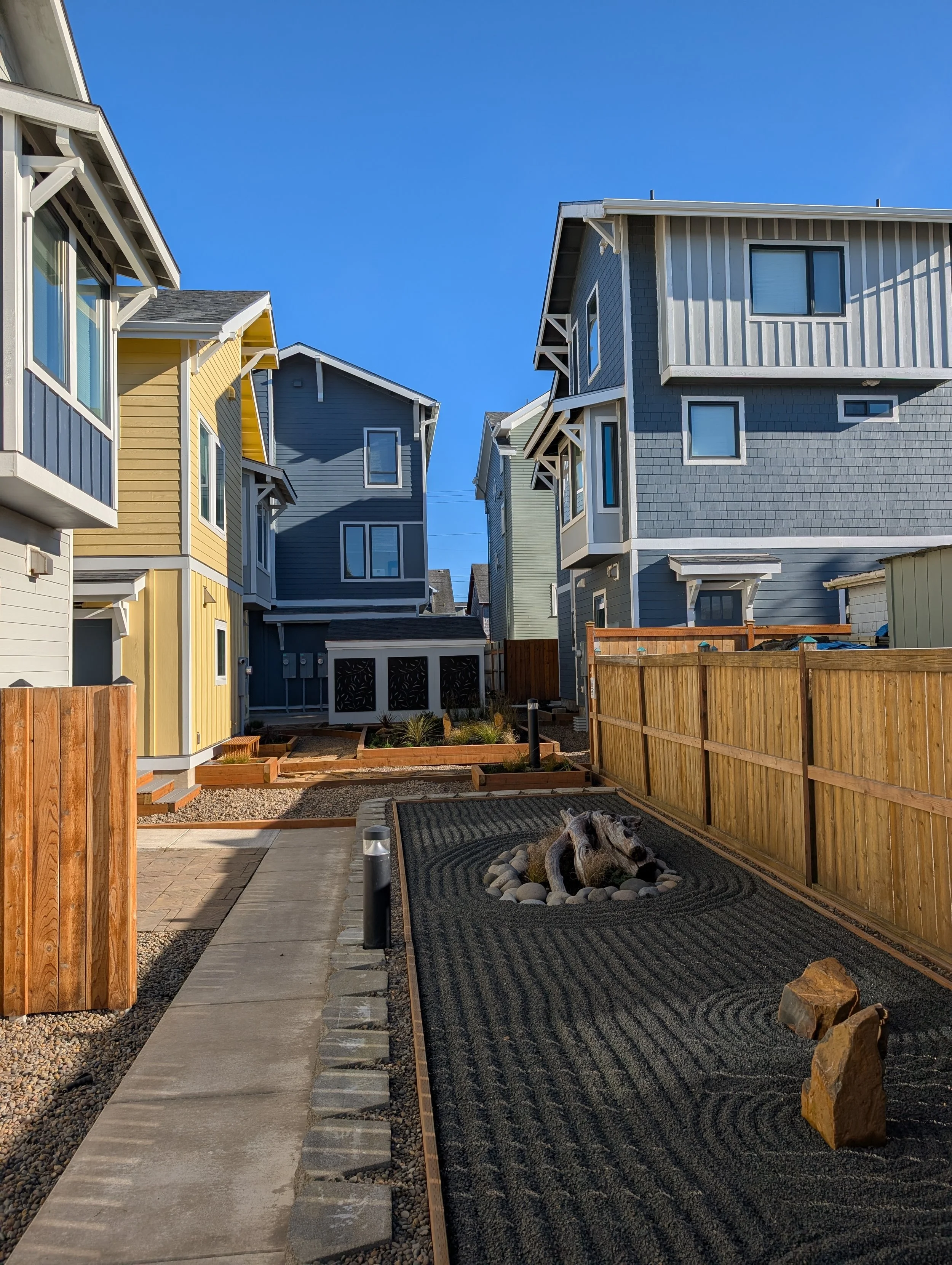 Side yard of colorful modern houses with fencing, landscaped rocks forming zen rake garden, and garden art of a dog sculpture.