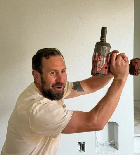 Man with short brown hair and short beard, wearing a white t shirt, makes a silly face while posing with a drill