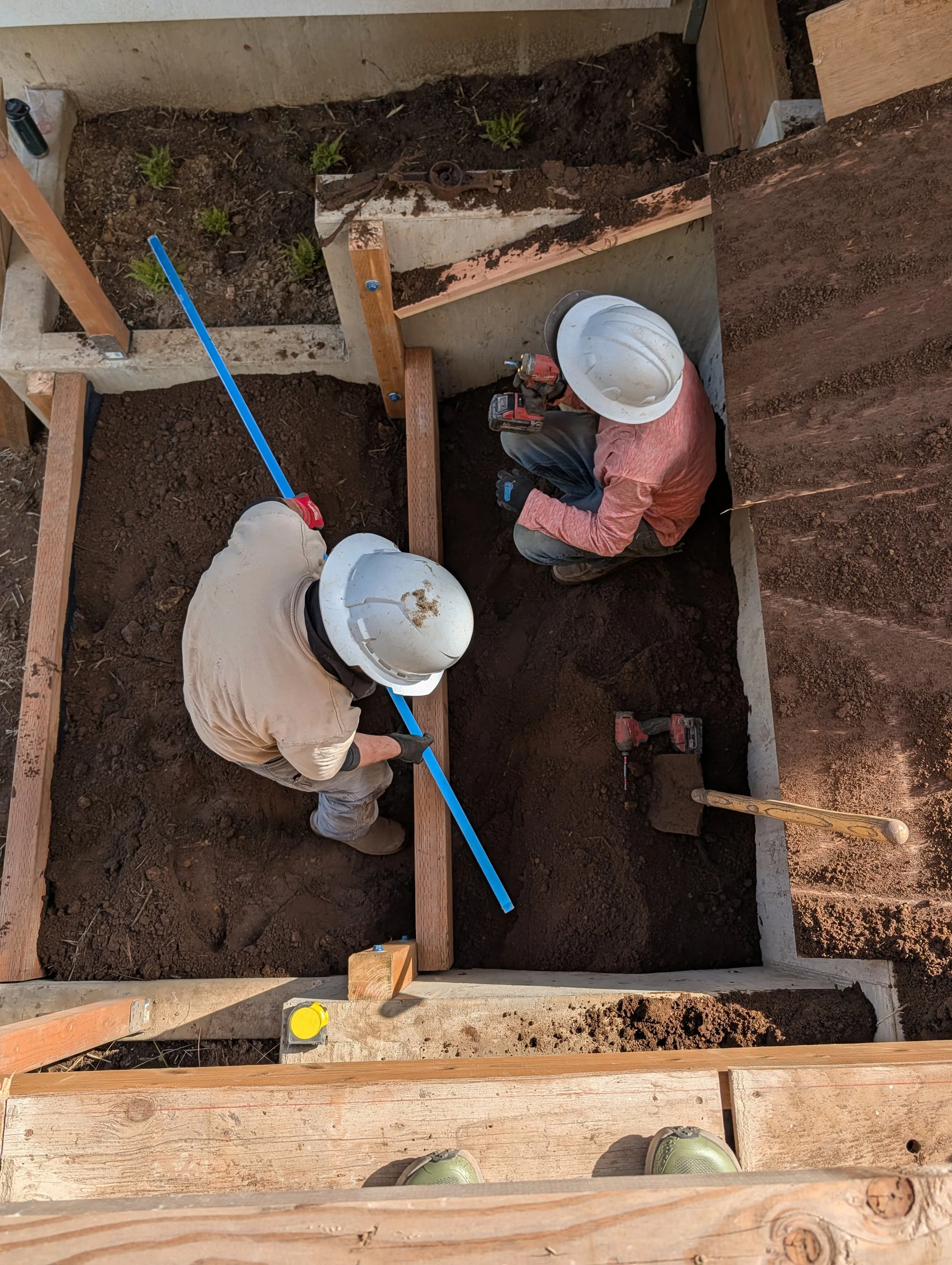 Two workers in construction hardhats working in a small outdoor dug-out area, installing or repairing a foundation with soil. Various tools and wooden framing are visible around the work area.