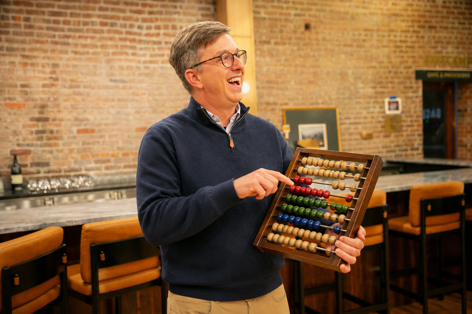 A man with glasses wearing a navy sweater smiling and holding a colorful abacus in a cozy, rustic bar or restaurant with brick walls and wooden tables.