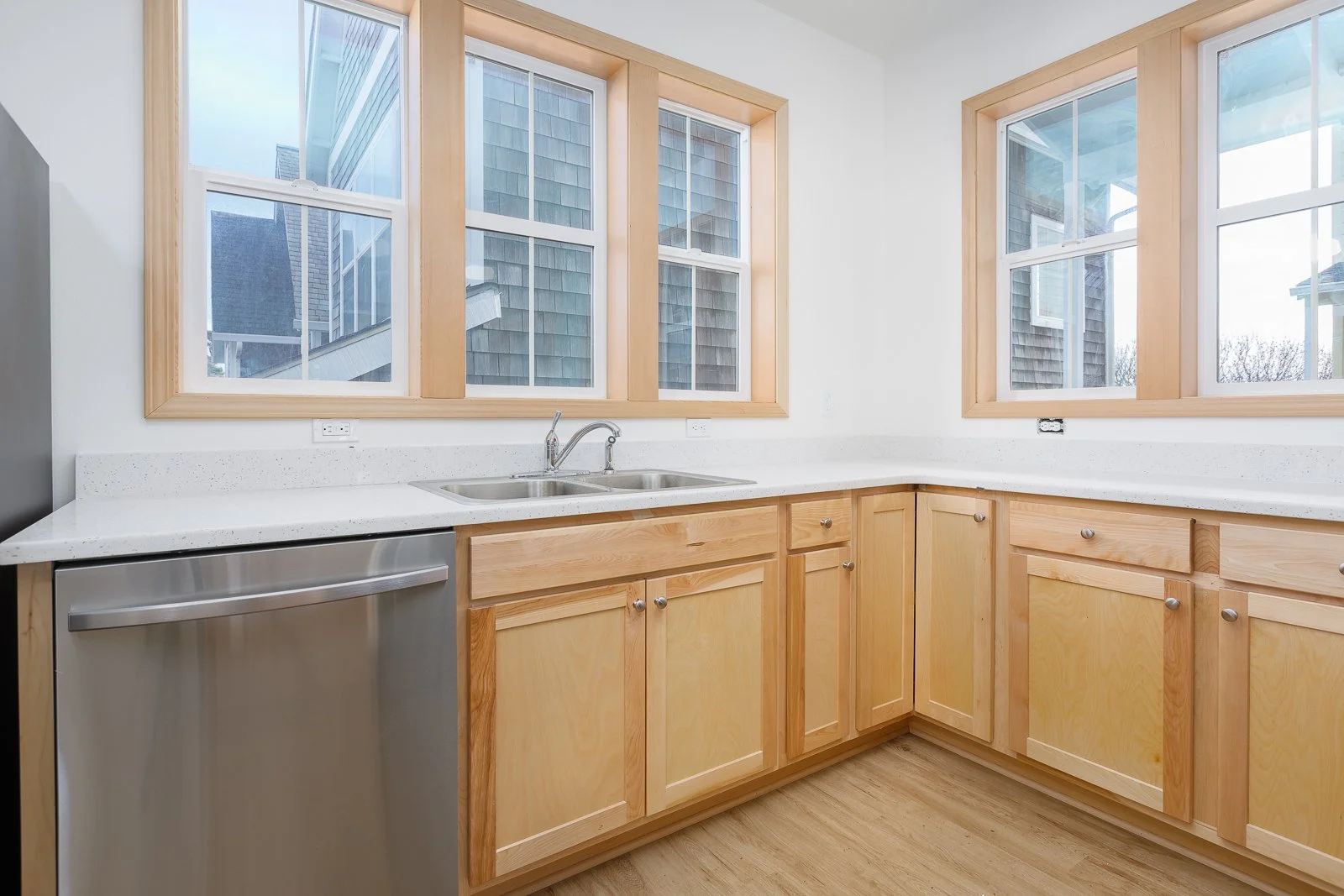 Kitchen with warm colored wooden cabinets, white countertops, a double sink, and large windows showing the exterior of neighboring houses.