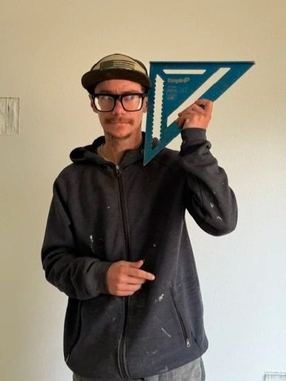 Man with large thick glasses wearing a black zip up jacket and a baseball cap makes a silly face while holding a framing square to his cheek.