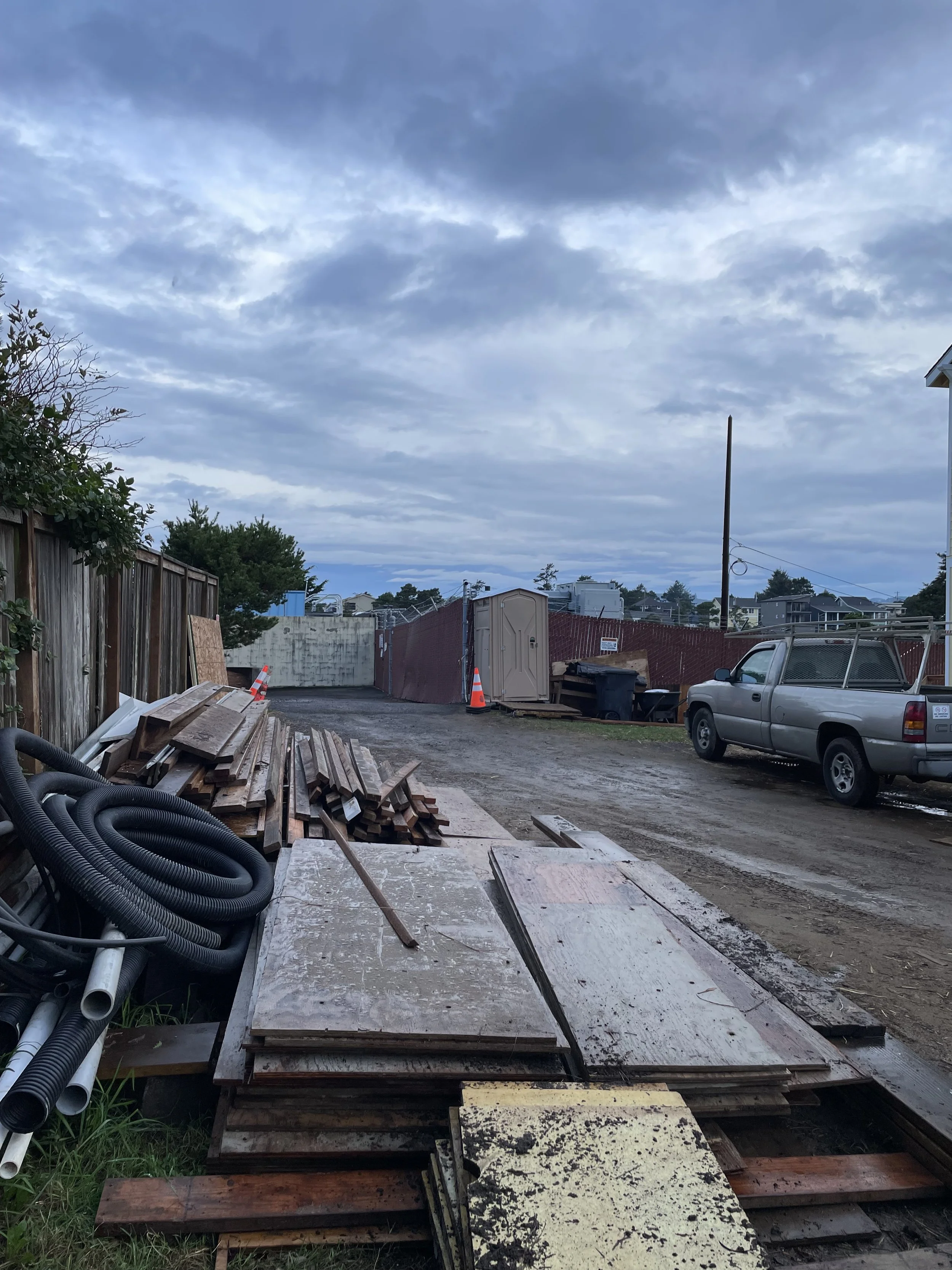 A well organized and orderly trash pile on a construction site. Featureing an overcast sky, a port-o-john, and a work truck.