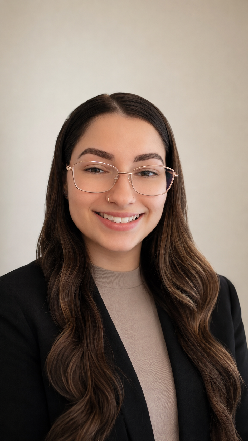 A young woman with long wavy brown hair, wearing glasses with a gold frame, a beige top, and a black blazer, smiling in front of a plain light-colored wall.