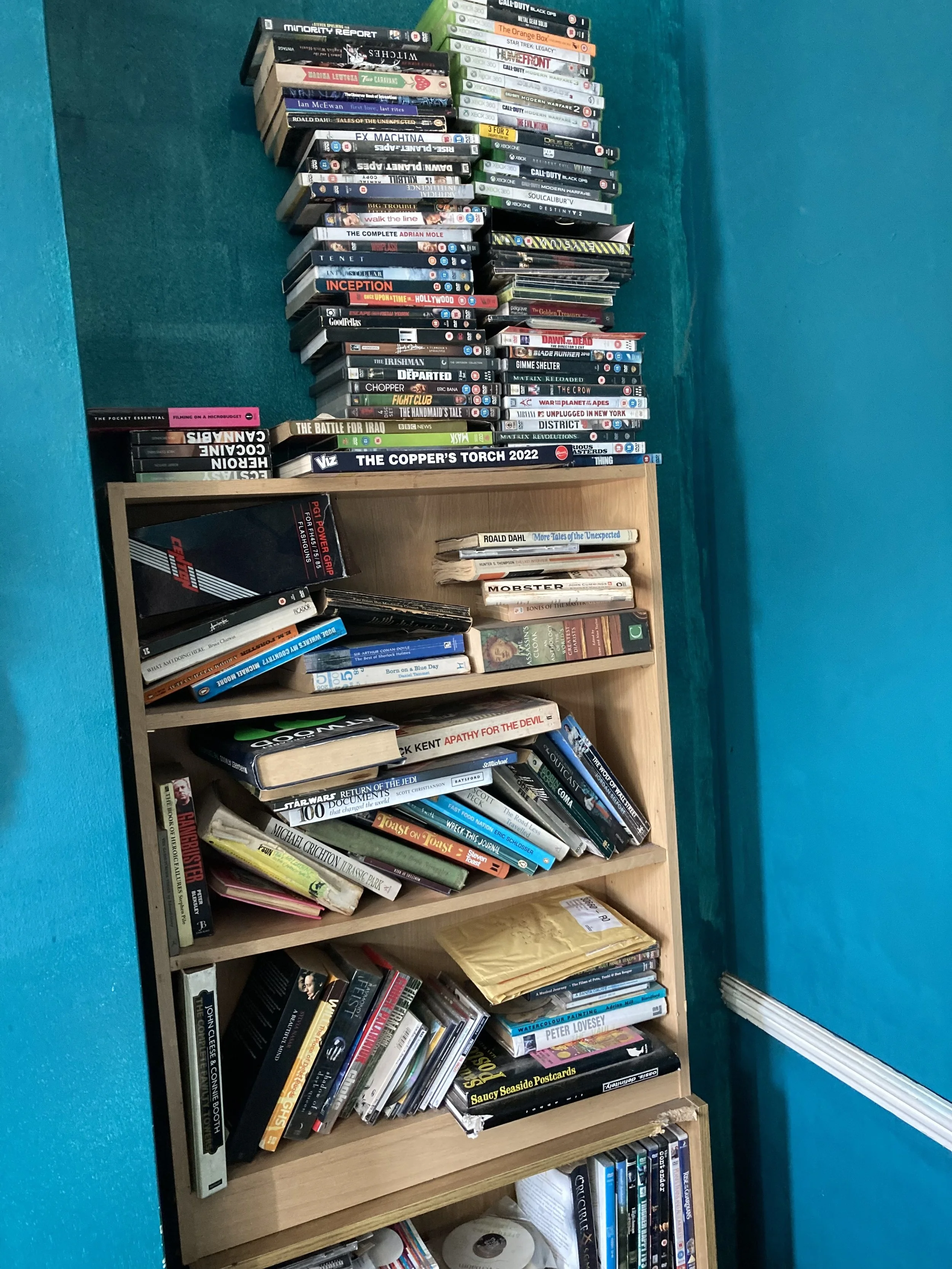 A wooden bookshelf filled with books and DVDs against a blue wall. The top of the shelf holds a stack of DVDs, including titles like 'Call of Duty,' 'Star Trek,' and 'Modern Warfare.' The middle shelves contain books such as 'The Battle for Iraq' and