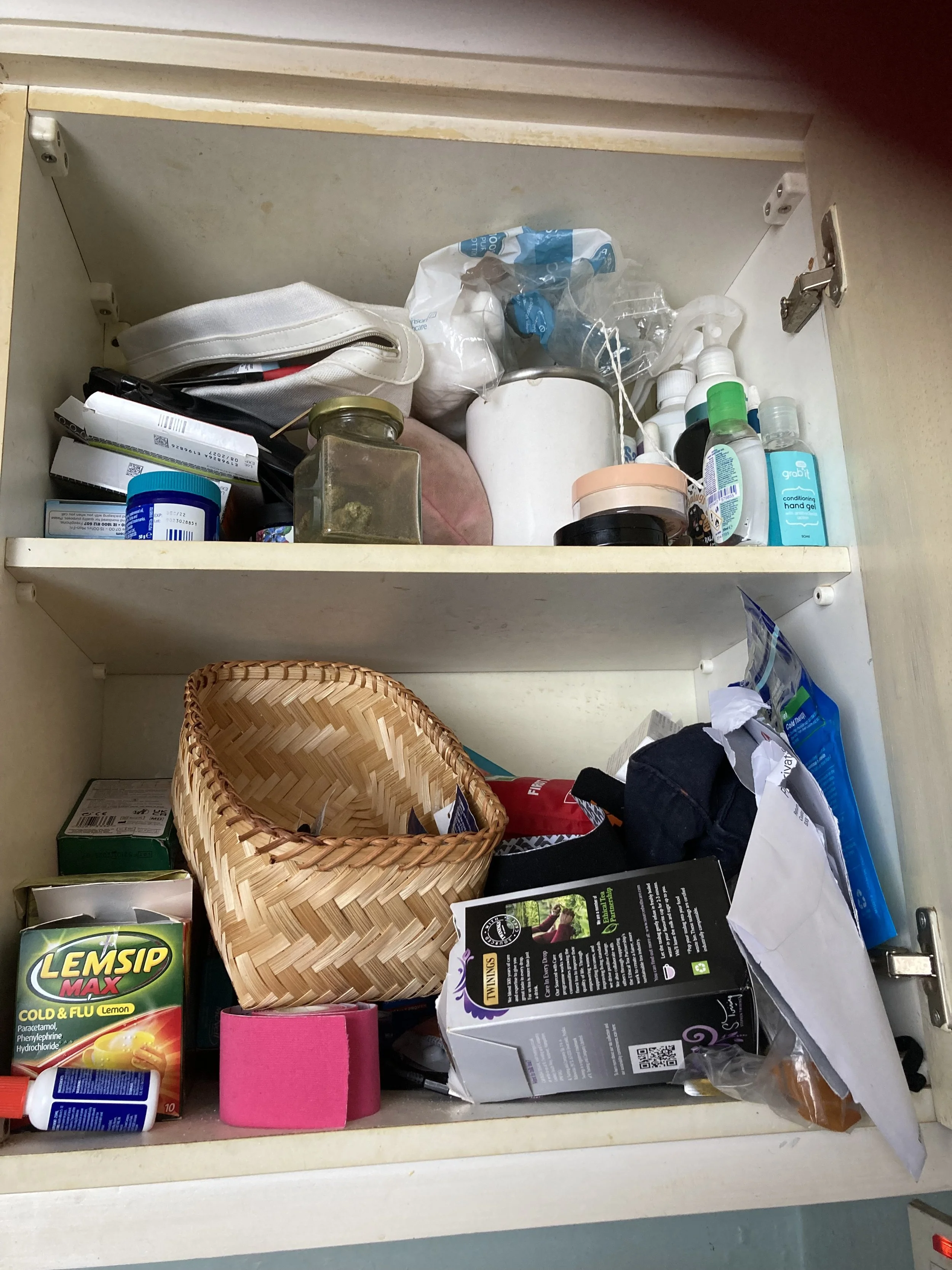 Open kitchen drawer containing various items including a wicker basket, medication boxes, a pink tape, a box of LemSip cold and flu lemon medicine, a tube of hand sanitizer, various bottles, a plastic bag, a pouch, and scattered papers.