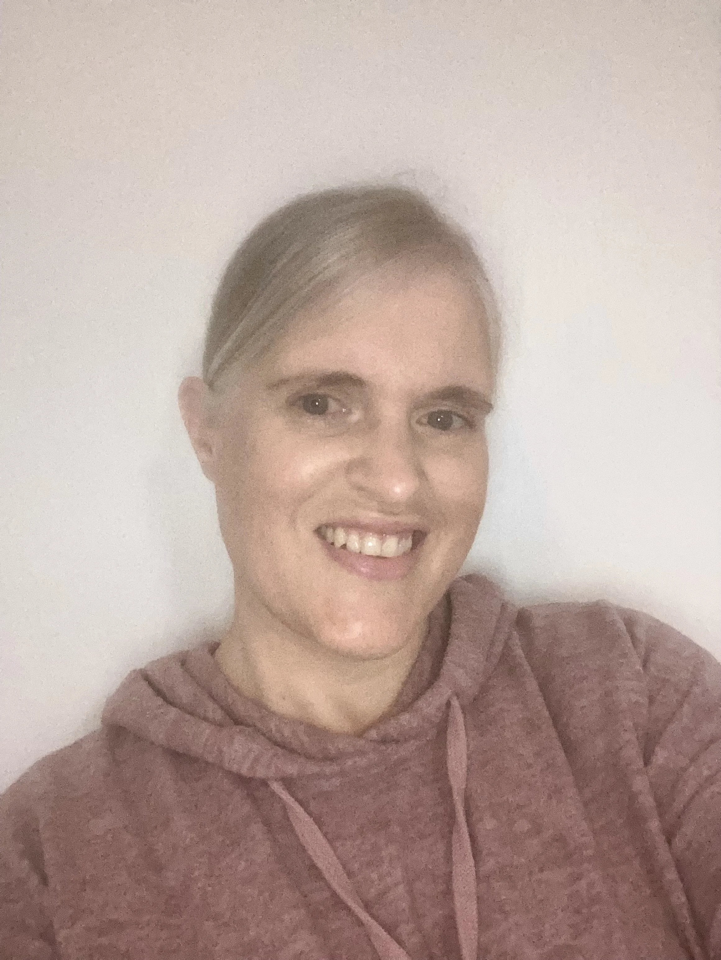 A woman with short blonde hair smiling, wearing a maroon hoodie, in front of a plain indoor wall.