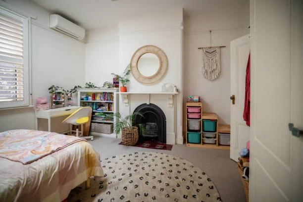A cozy bedroom with a bed, a white desk and yellow chair, a bookshelf, a fireplace, a round mirror, a plant, a woven wall hanging, and colorful storage bins.