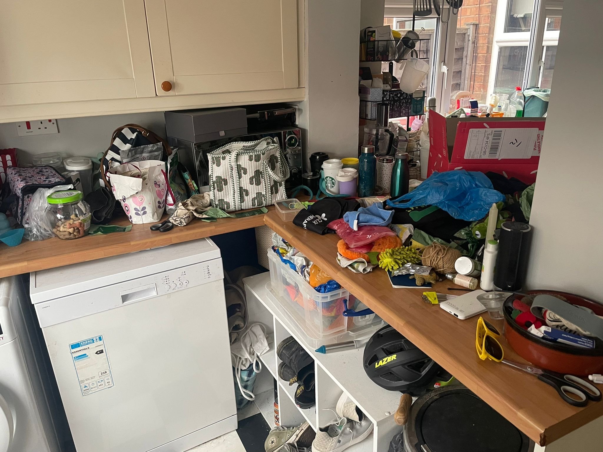 Cluttered kitchen worktop with various items including reusable Starbucks cups, bags, scissors, gloves, and cleaning supplies, with cabinets and a window in the background.