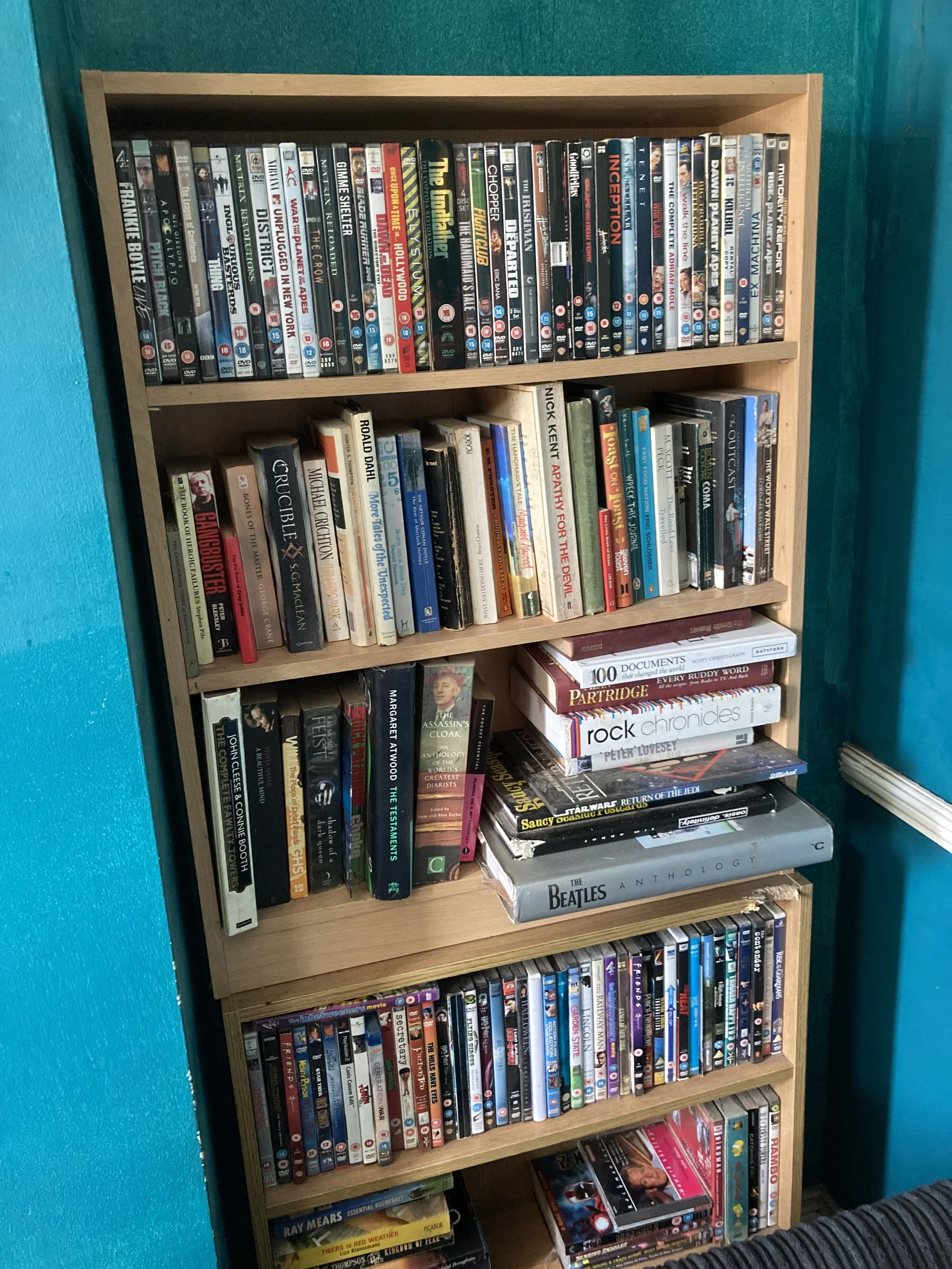 A wooden bookshelf filled with movies, books, and DVDs, placed against a teal wall.