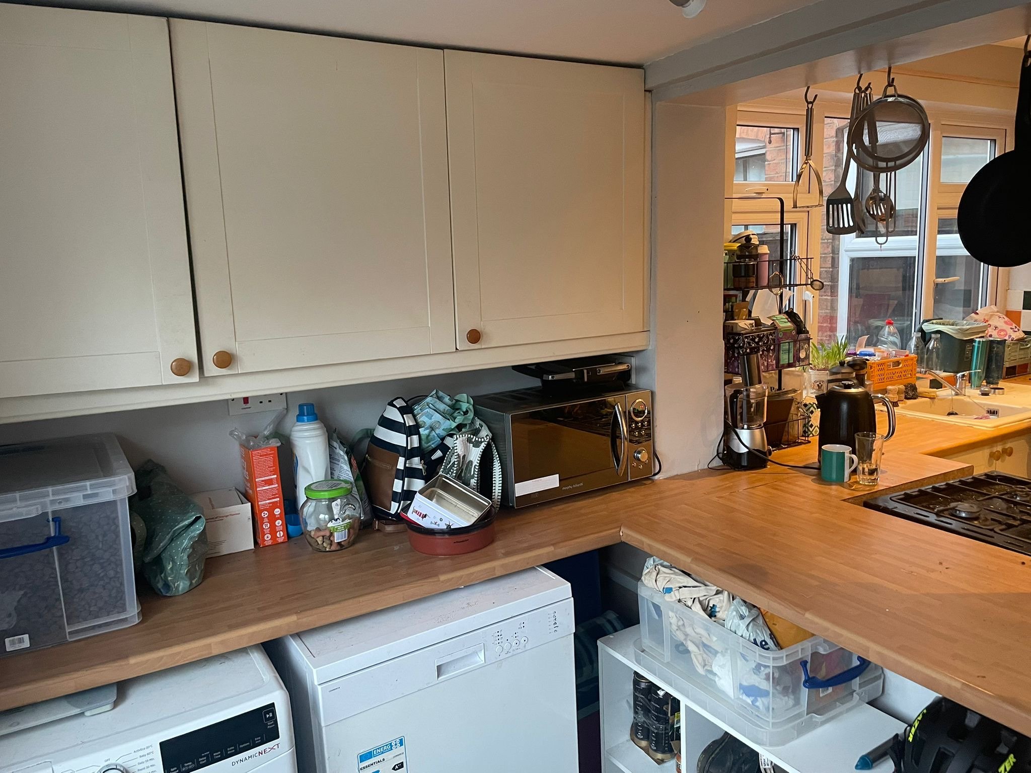 A kitchen with white cabinets, a wooden countertop, and various kitchen utensils and appliances. There are laundry machines underneath the counter and a window with a view outside.