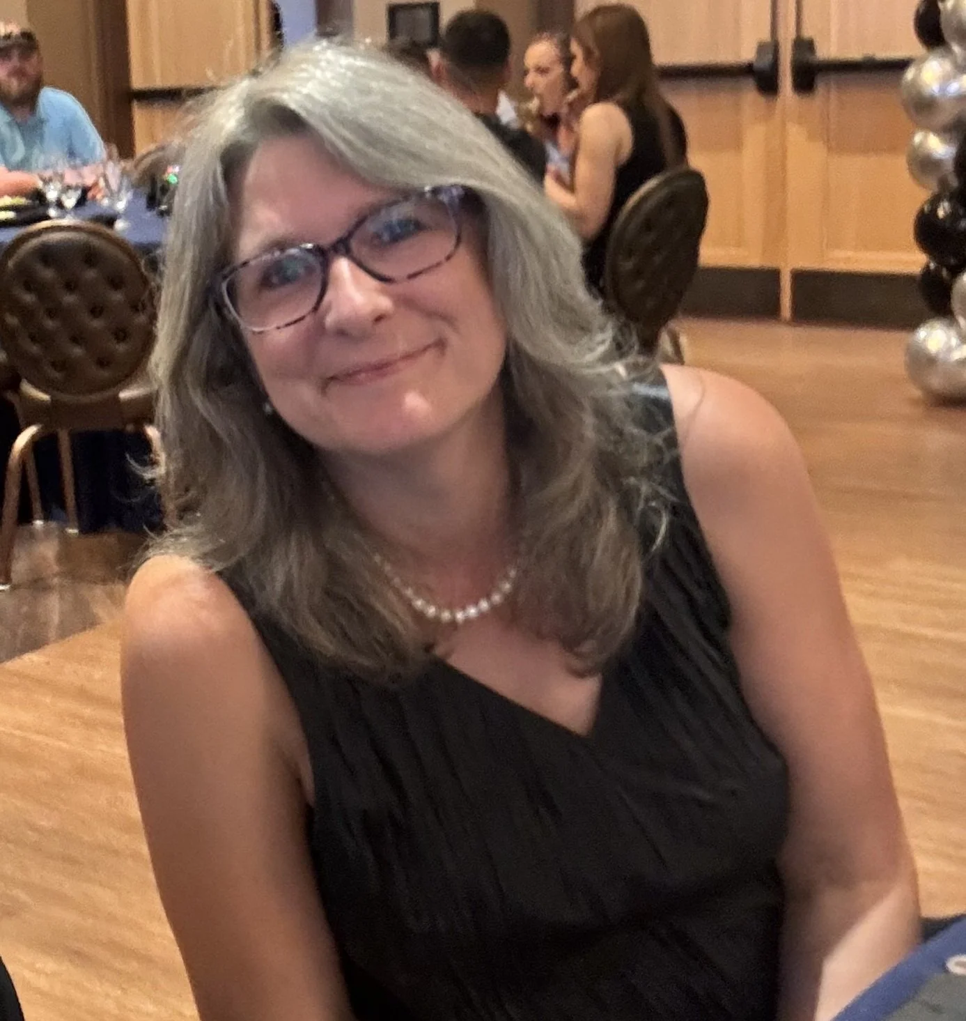 A woman with gray hair, glasses, and a pearl necklace, smiling at a social gathering in a restaurant or banquet hall.