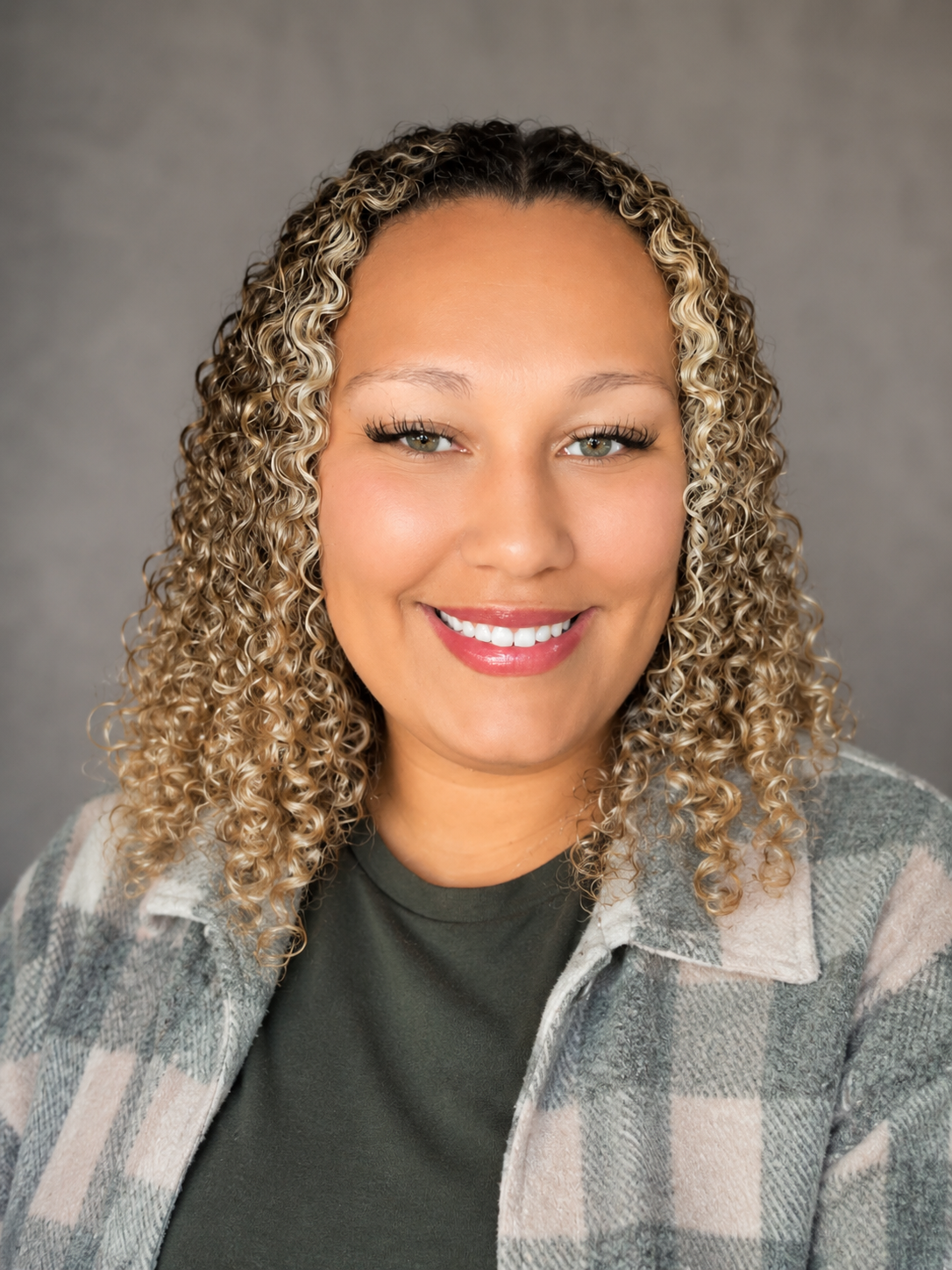 Portrait of a woman with curly blonde hair, smiling, wearing a gray and beige plaid shirt over a dark green top, against a gray background.