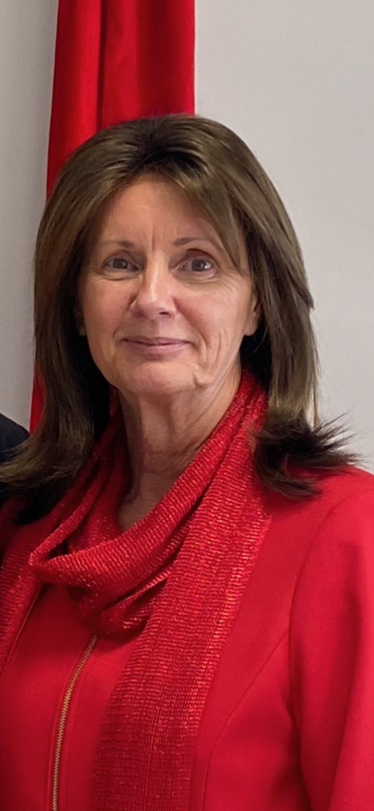 A woman with brown hair, wearing a red blazer and scarf, standing in front of a red and white background.