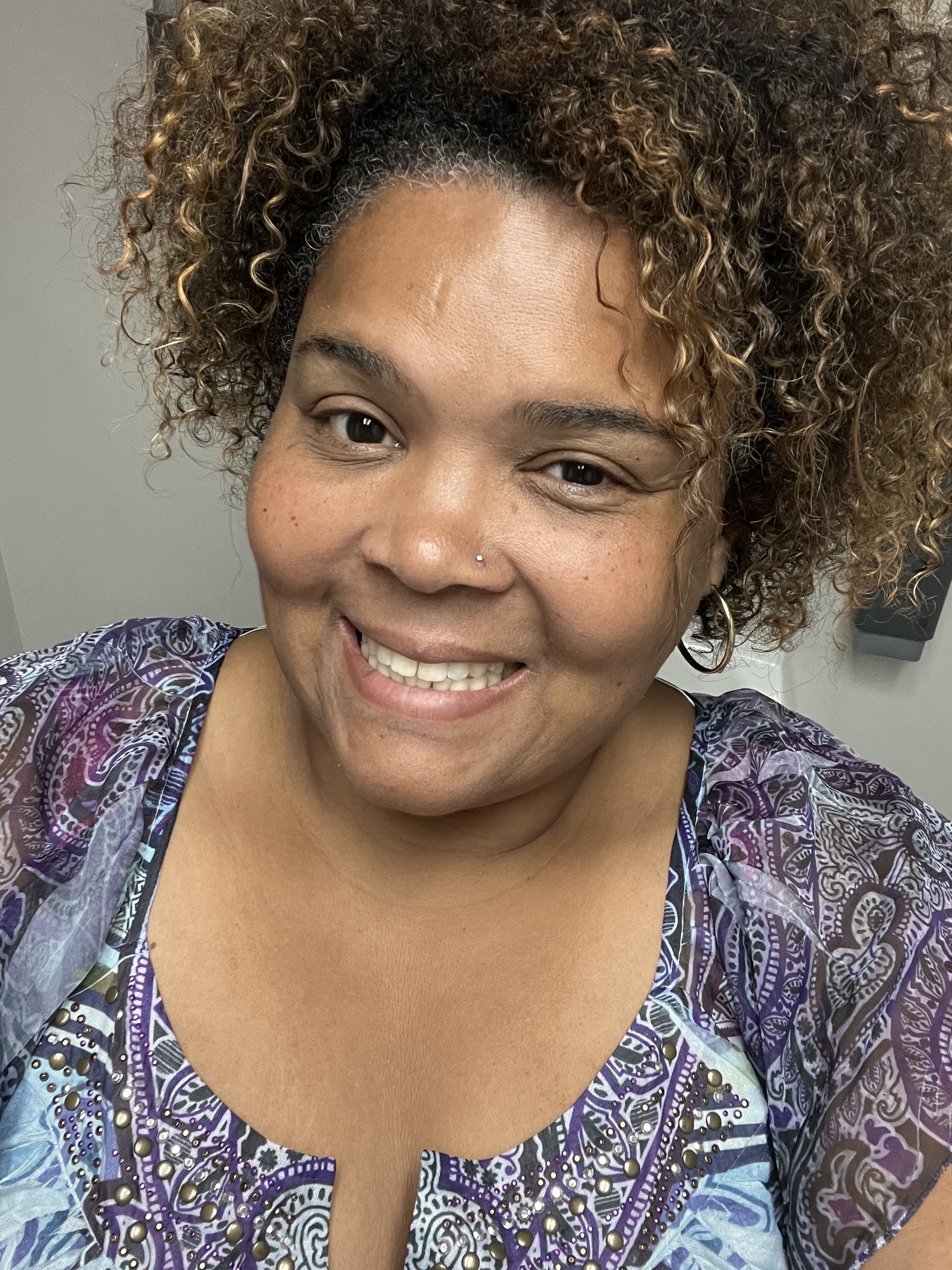 A woman with medium skin tone, curly brown hair with golden highlights, smiling and showing her teeth, wearing a purple patterned top with metallic embellishments.