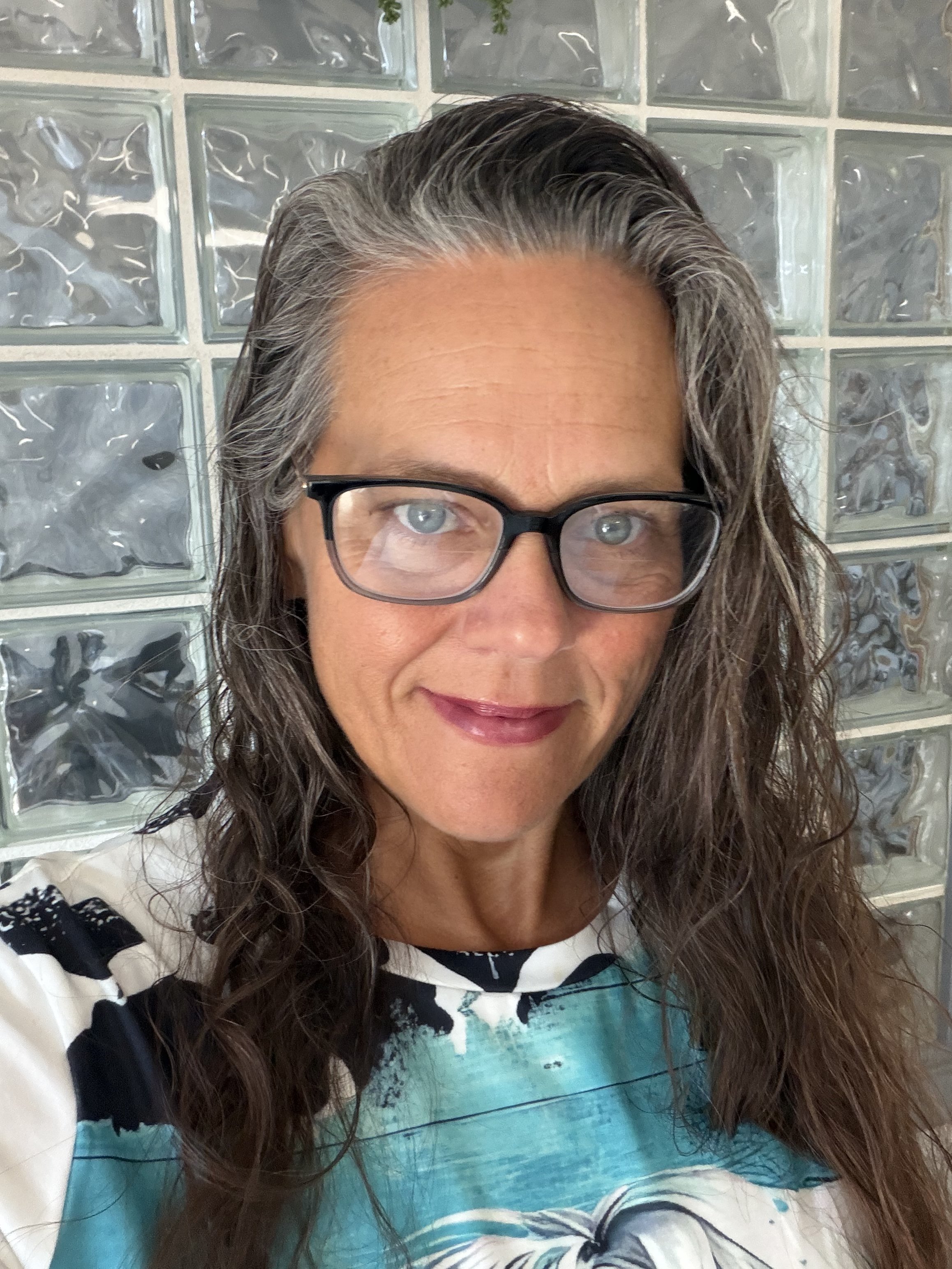 A woman with shoulder-length gray and brown wavy hair and glasses smiling at the camera. She is in front of a glass block wall and wearing a white top with a colorful print.