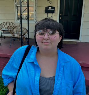 A young woman with short dark hair and glasses, wearing a bright blue jacket over a gray shirt, standing outside a house with a black door and porch.