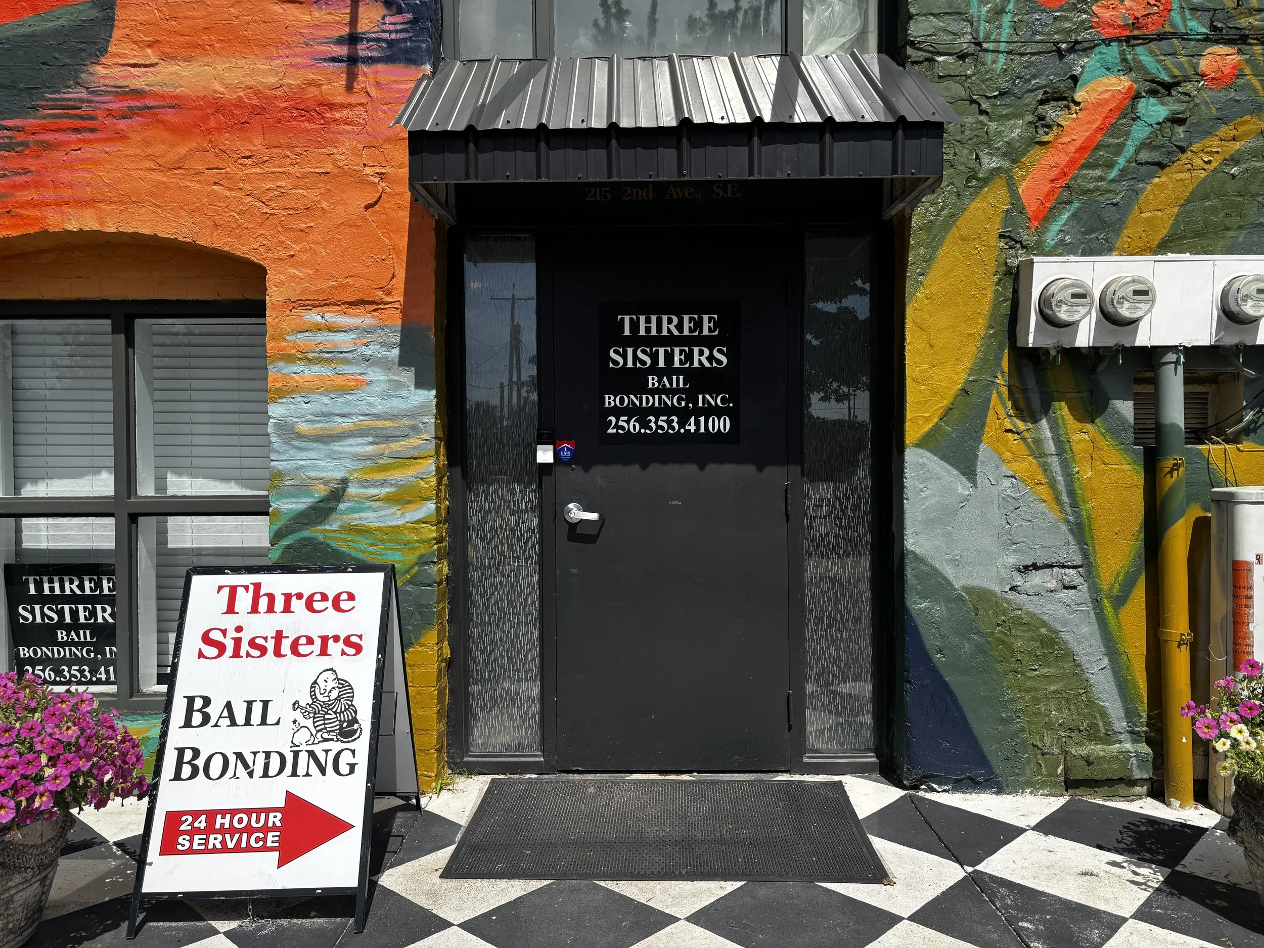 The entrance to a bail bond business called 'Three Sisters Bail Bonding, Inc.' with a black door, colorful mural painted on the wall, potted flowers, and a sign indicating 24-hour service.