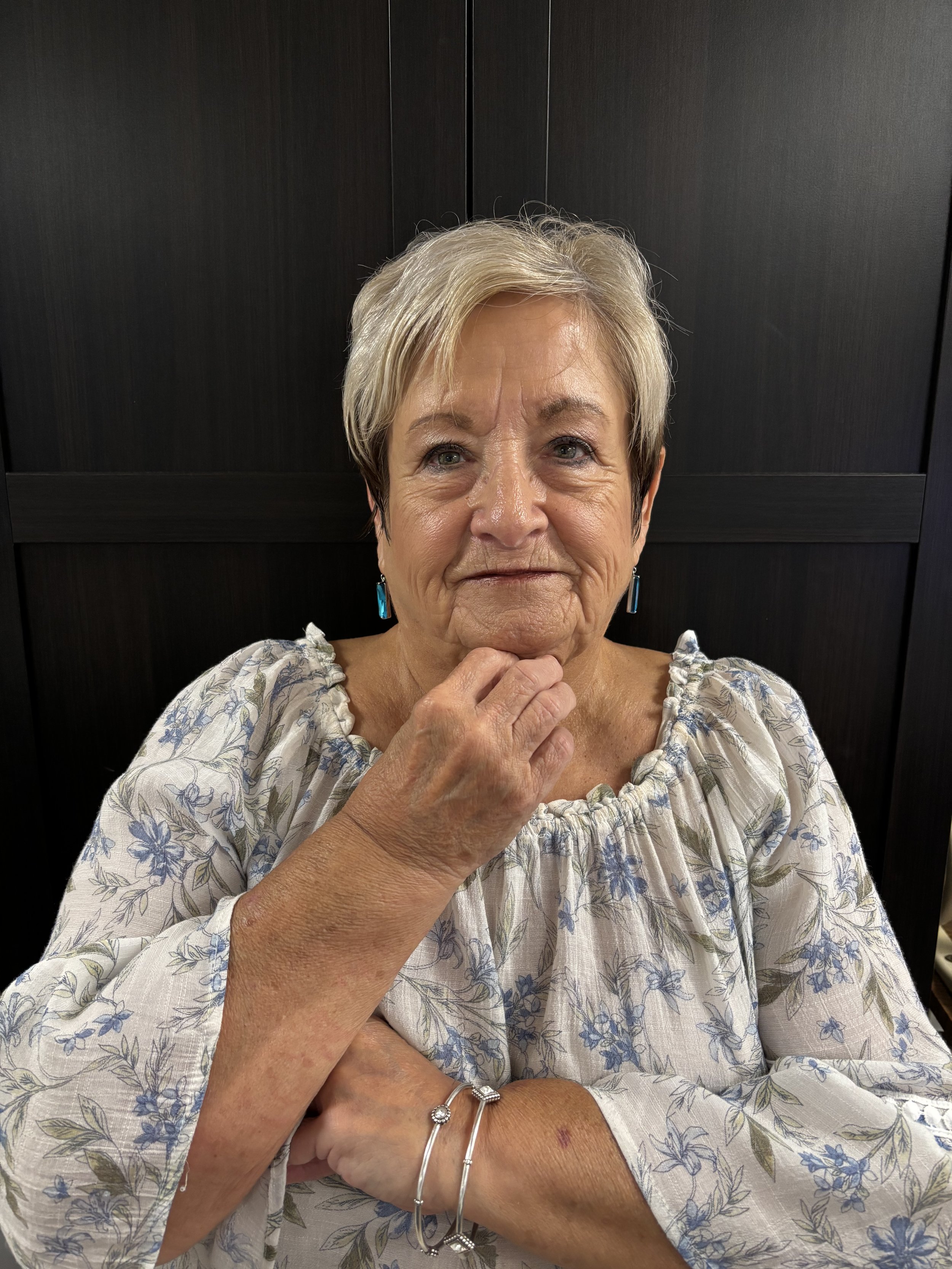 An elderly woman with short gray hair, wearing a floral patterned dress and blue earrings, poses with her hand under her chin in front of a dark wooden panel background.