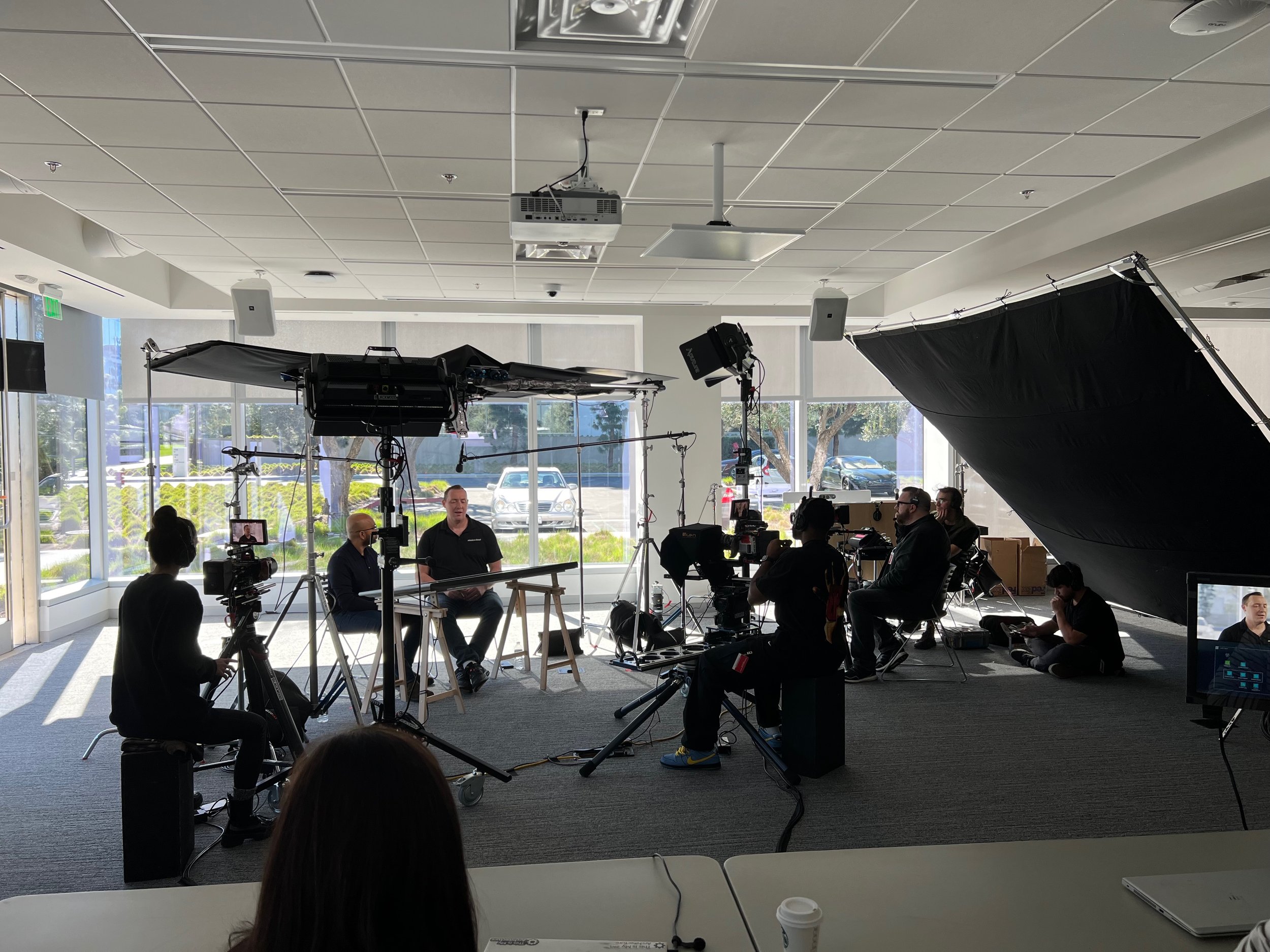 People filming a video interview or presentation in a bright room with large windows. Camera and lighting equipment are visible, with some crew members operating them, while two men sit at a table being filmed.