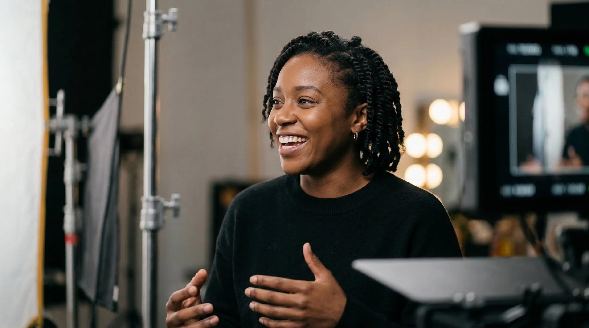 A woman smiling and touching her chest in a studio setting, with professional lighting and equipment in the background.