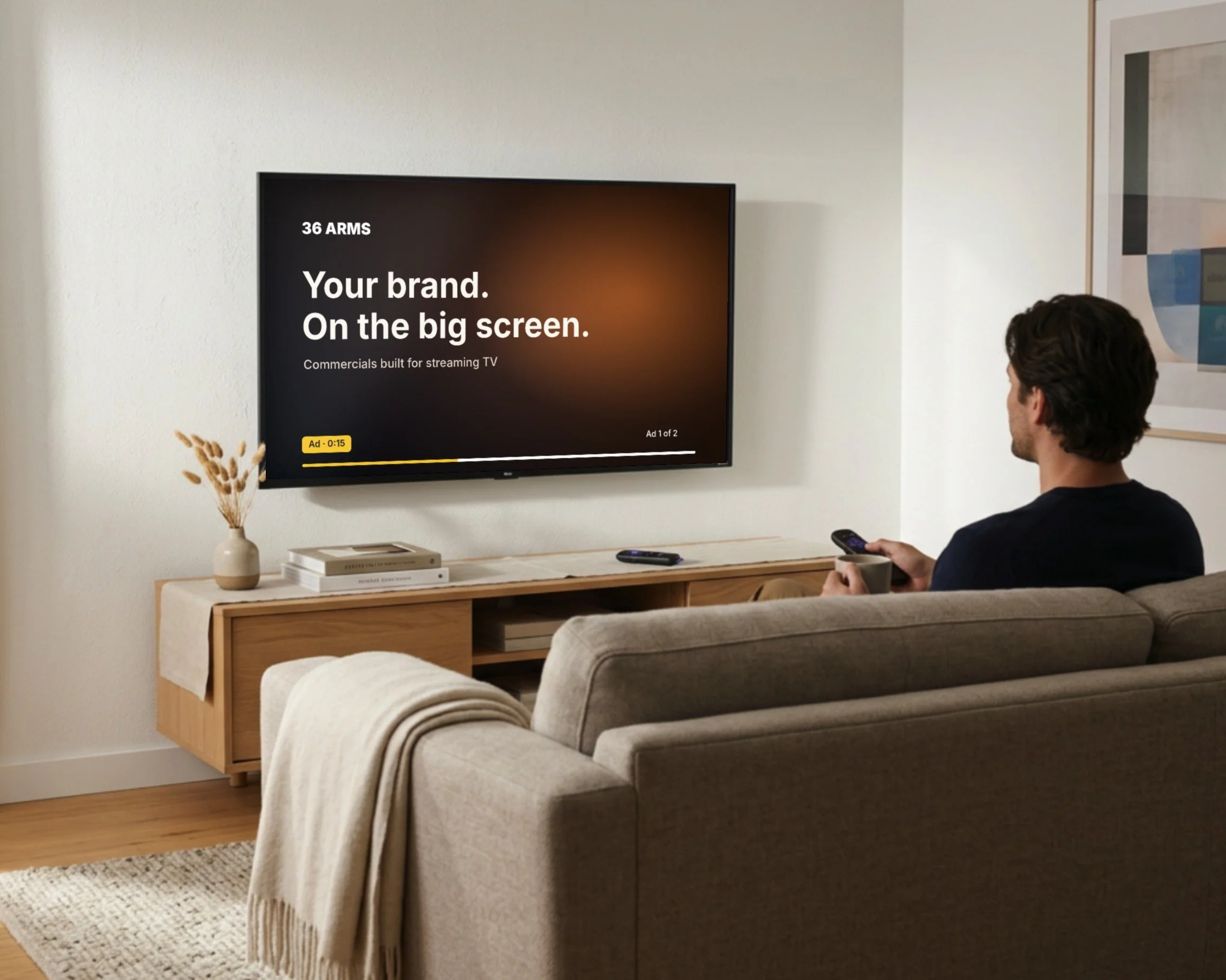 Living room with a beige sofa, a wooden TV stand with books and a vase, a flat-screen TV displaying an advertisement, and a man sitting on the sofa holding a remote control.