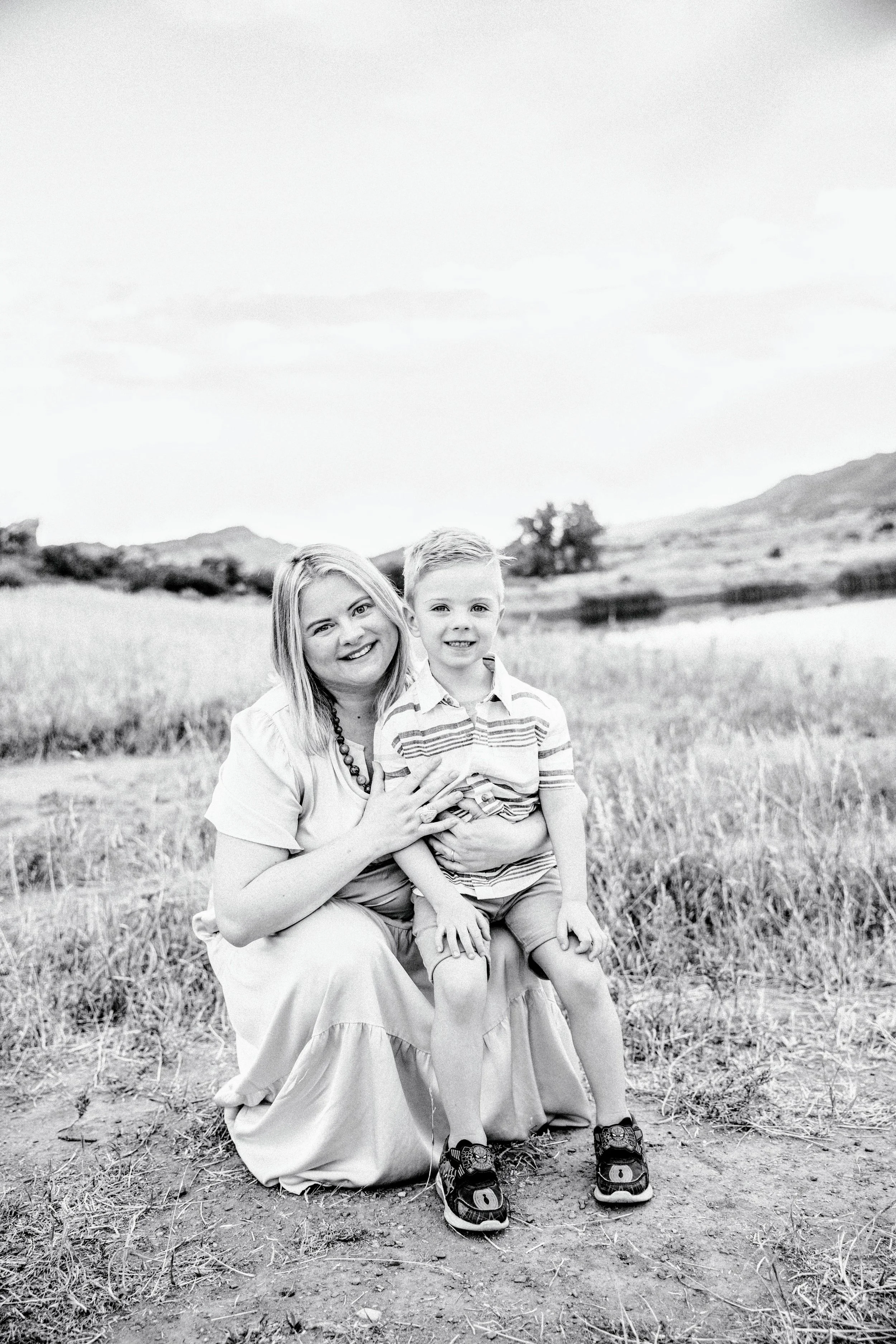 A black and white photo of a woman with medium-length hair and a young boy outdoors in a grassy area with mountains in the background. The woman is kneeling on one knee, smiling, and holding the boy who is sitting on her knee, also smiling. The boy is wearing a striped polo shirt, shorts, and sneakers.
