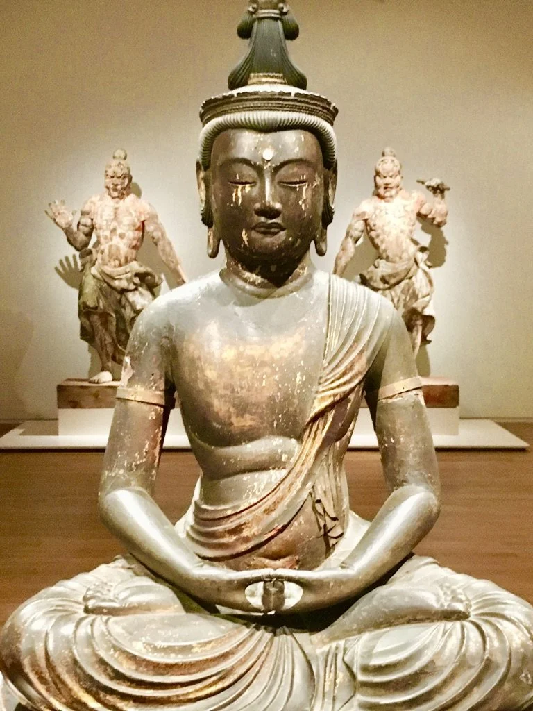 Ancient seated Buddha sculpture with closed eyes, wearing a crown-like headdress, with two smaller standing sculptures in the background.